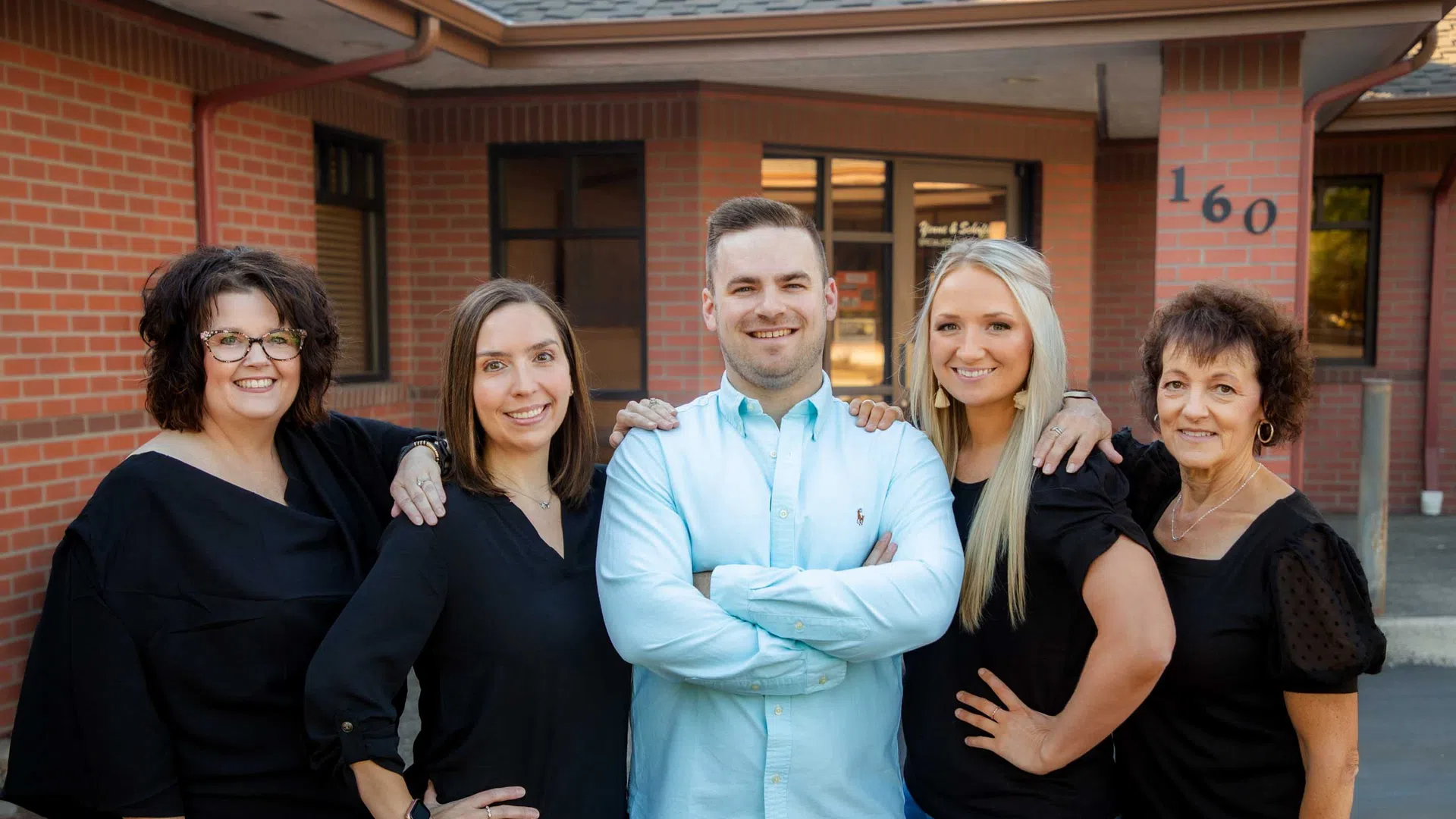 Five smiling adults standing confidently outside a brick building with address number 160 visible.