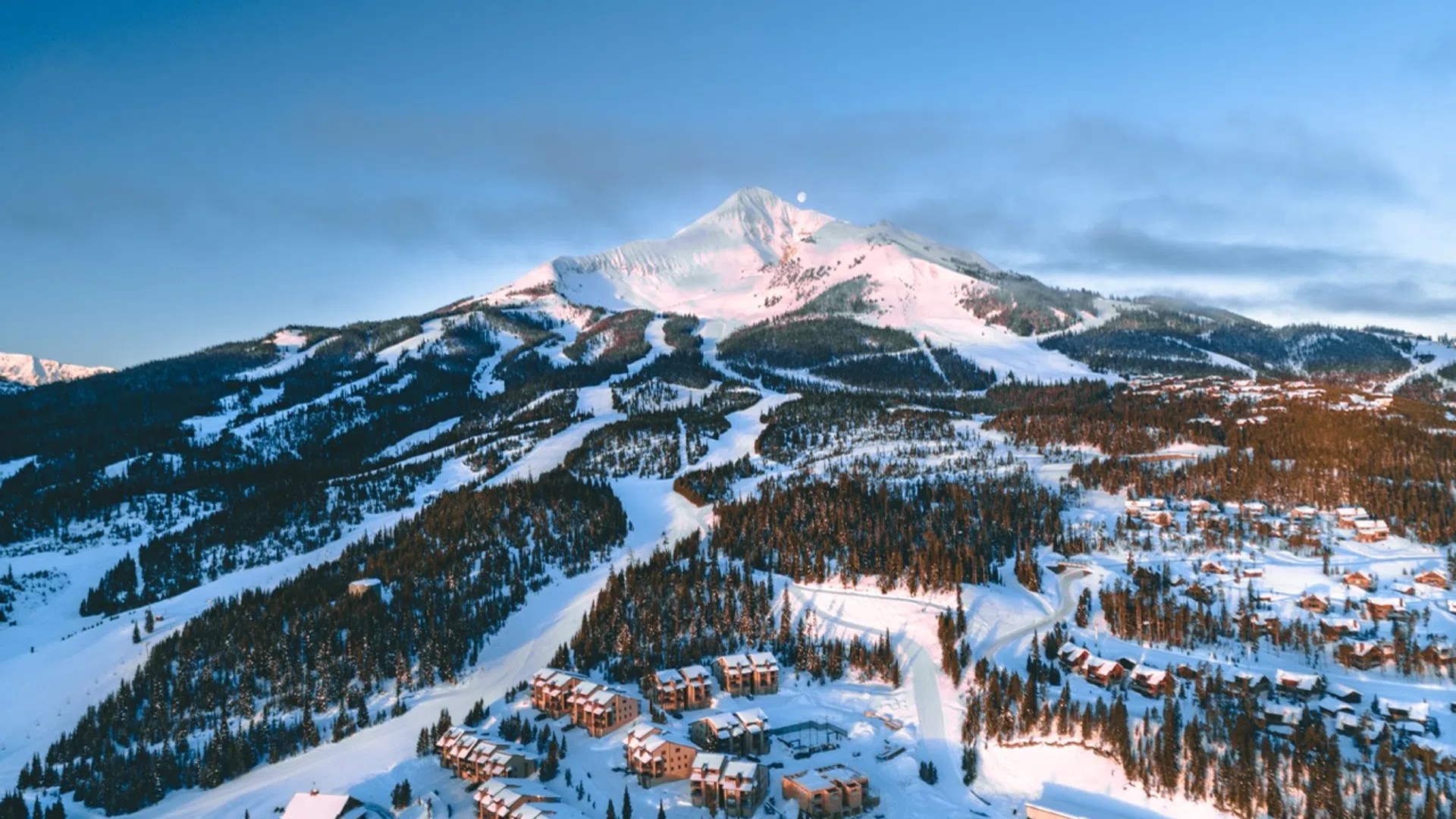 Snow-covered mountain with ski trails and resort buildings under a clear blue sky at sunset.