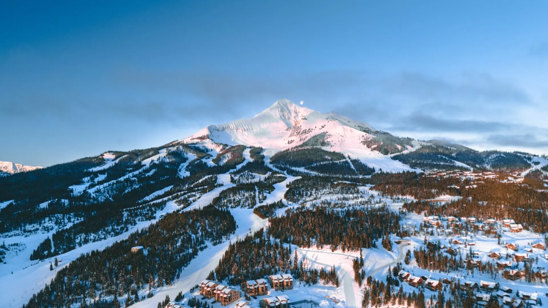 Snow-covered mountain with ski trails and resort buildings under a clear blue sky at sunset.
