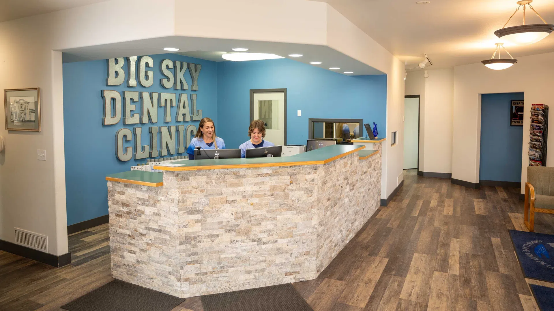 Reception area of Big Sky Dental Clinic with two staff members at a stone-themed front desk and blue accent wall.