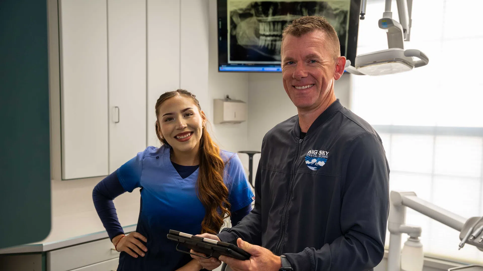 Smiling dental professionals in a clinic with an X-ray image on the monitor behind them.