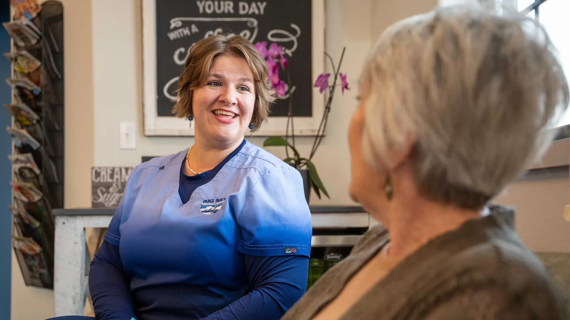 Smiling nurse in blue scrubs talking to elderly woman in a cozy indoor setting with plants and decor.