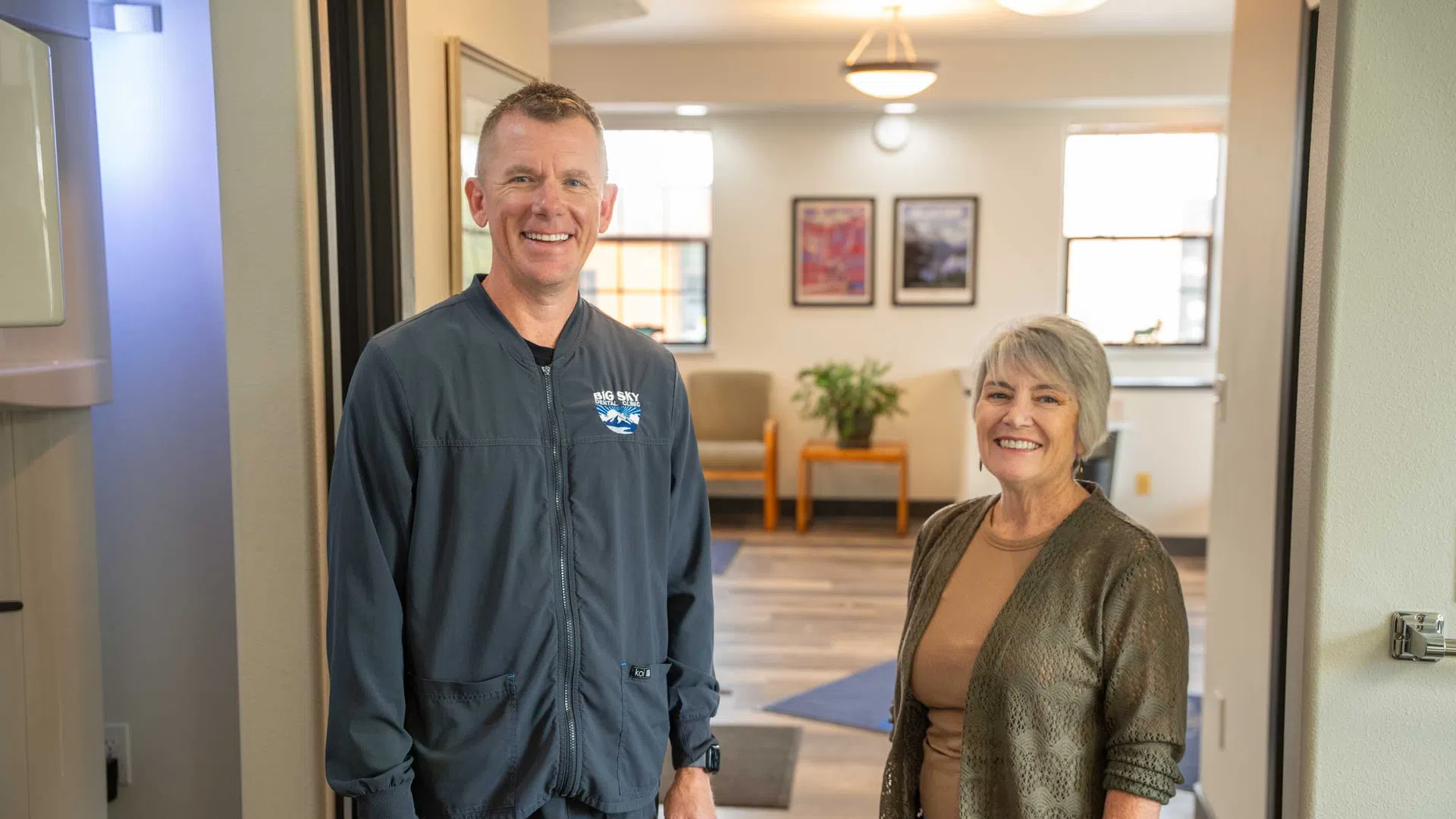 Smiling man in blue uniform and older woman standing in office hallway with chairs and plants in background
