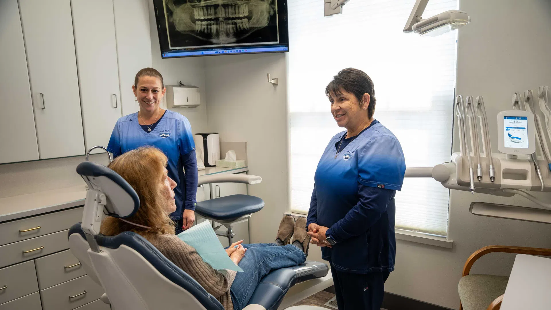 Dental patient in chair talking with two smiling dental hygienists in modern clinic with x-ray on screen.