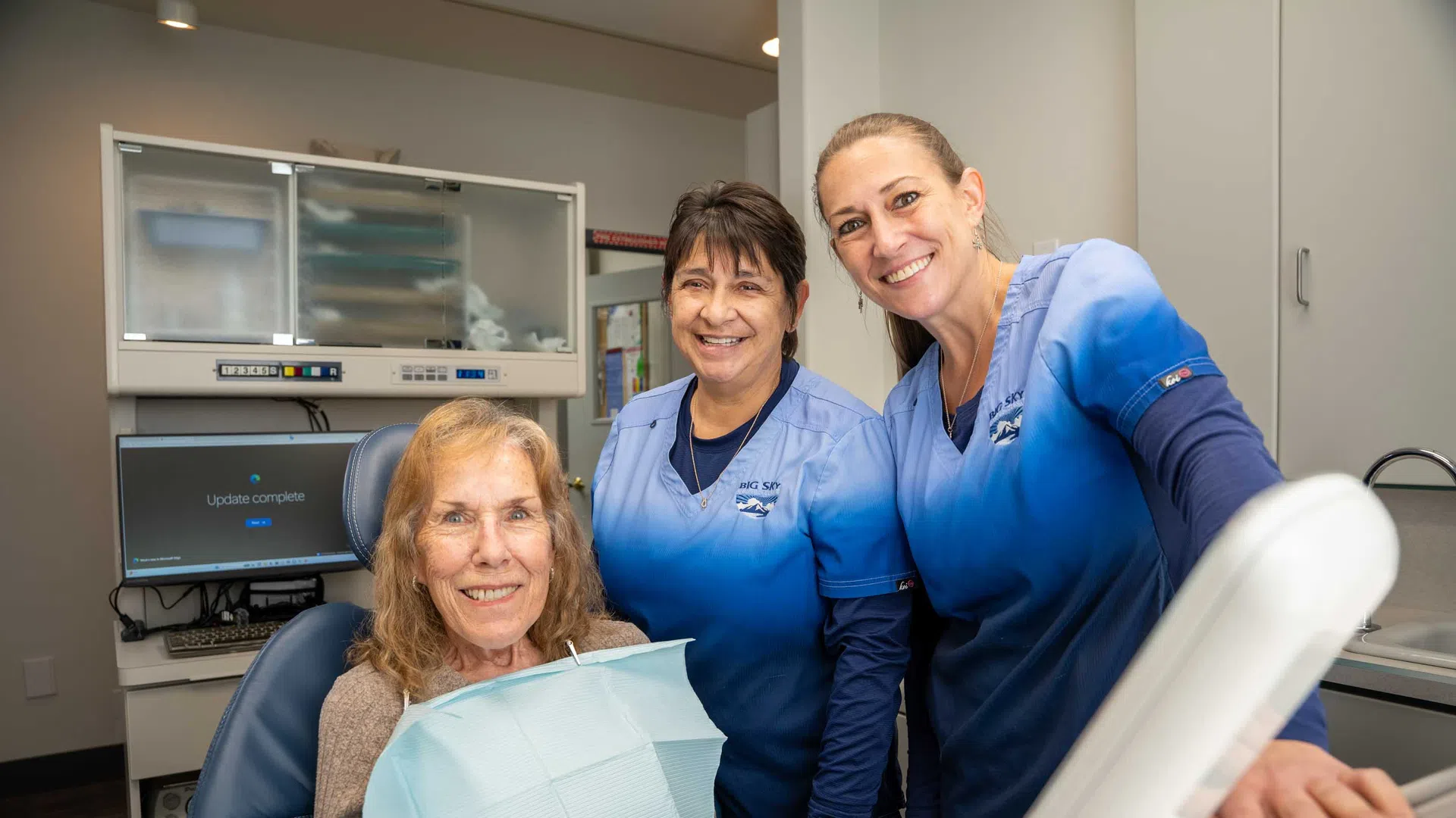 Smiling dental patient seated with two dental hygienists in blue scrubs in a modern clinic room.