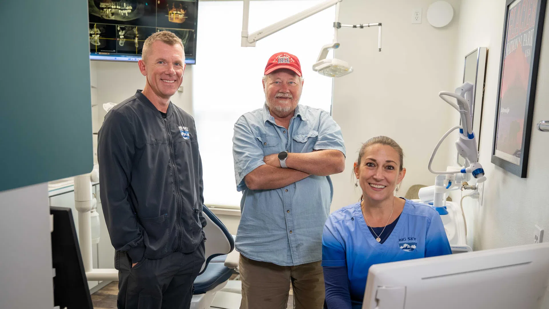 Two dental professionals and a patient smiling inside a modern dental clinic with equipment and computer screen visible.