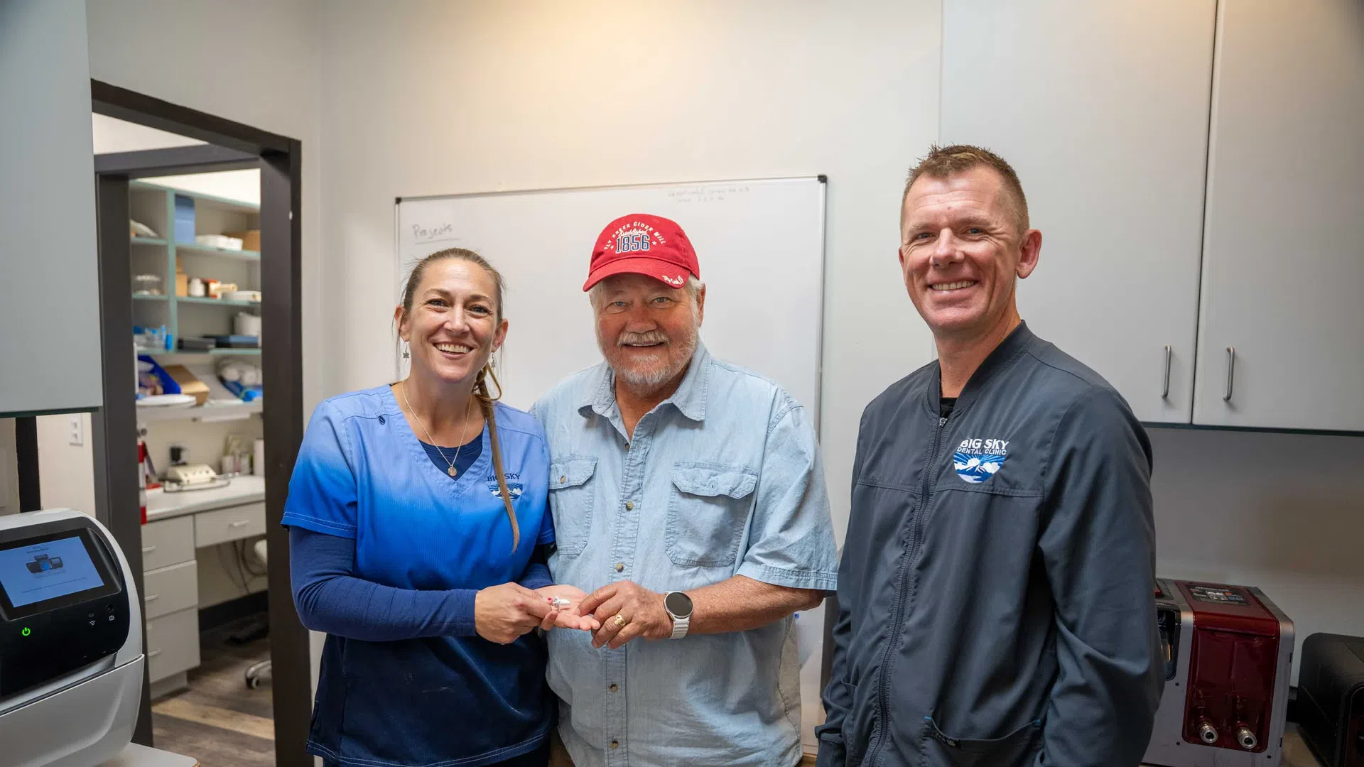 Smiling dental staff and patient in clinic room with equipment and cabinets, celebrating successful treatment.