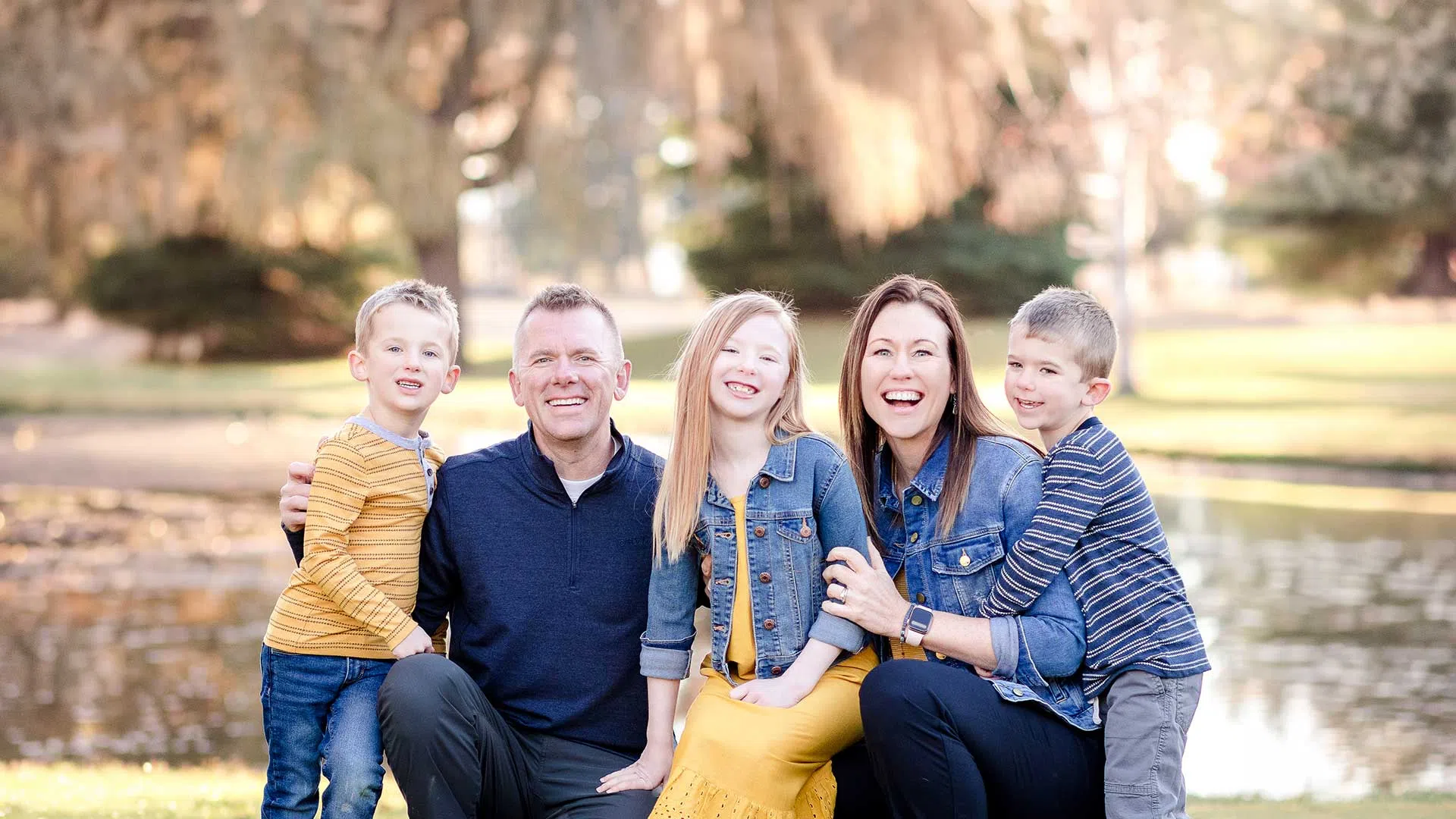 Happy family of five posing outdoors by a pond with trees in the background on a sunny day