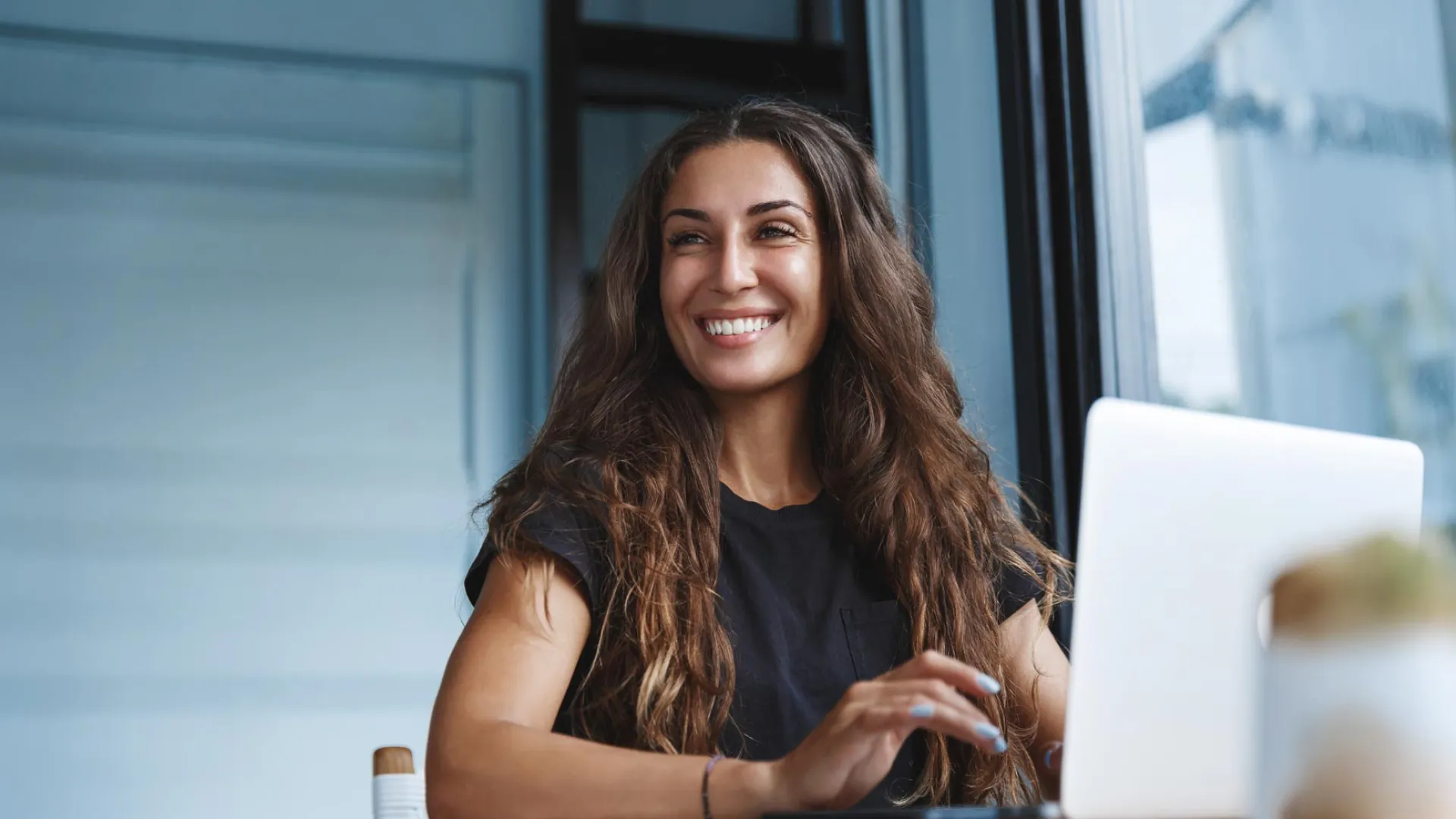 Smiling woman with long brown hair working on a laptop in a bright modern office space.