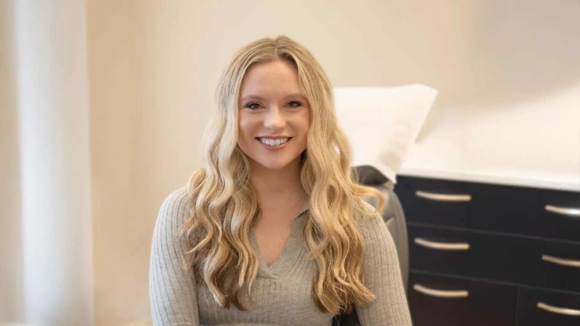 Smiling young woman with long wavy blonde hair sitting in a modern office chair in a bright room.