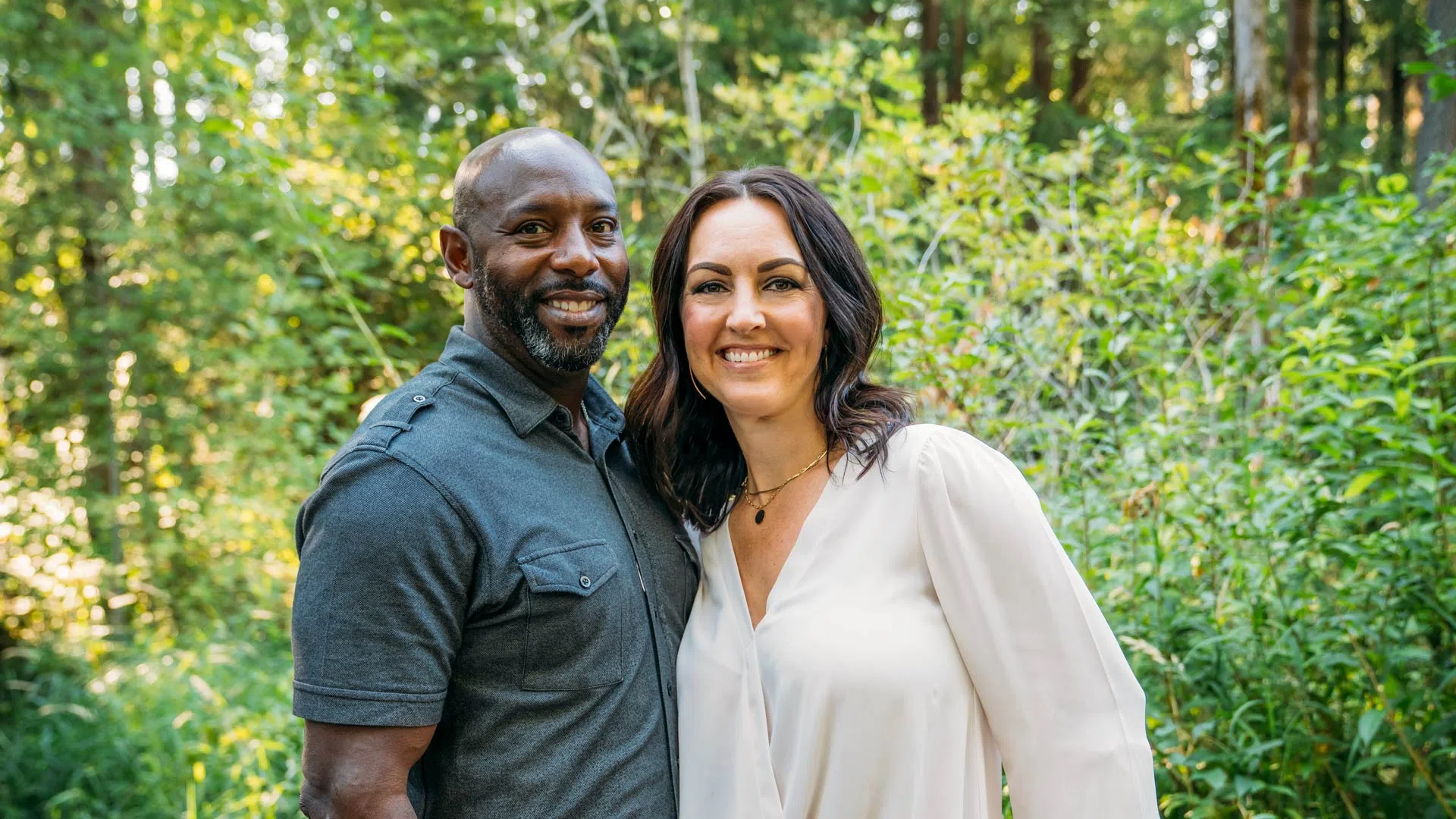 Smiling interracial couple standing close together outdoors with lush green forest background.