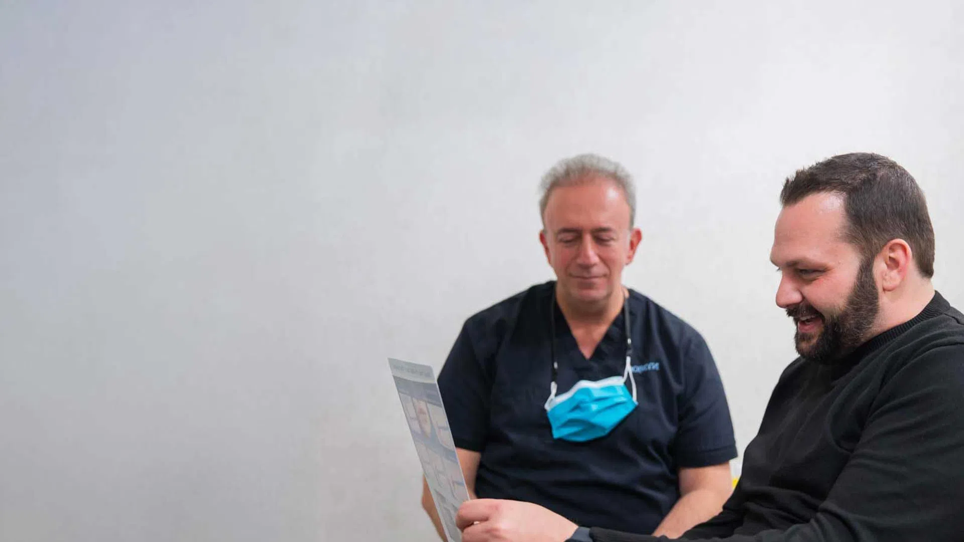 Two men, one in medical scrubs with a mask, reviewing a document together in a clinical setting.