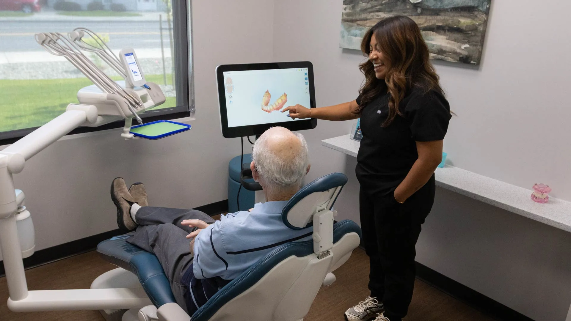 Dentist explaining dental scan results on screen to elderly patient reclining in dental chair in clinic room.