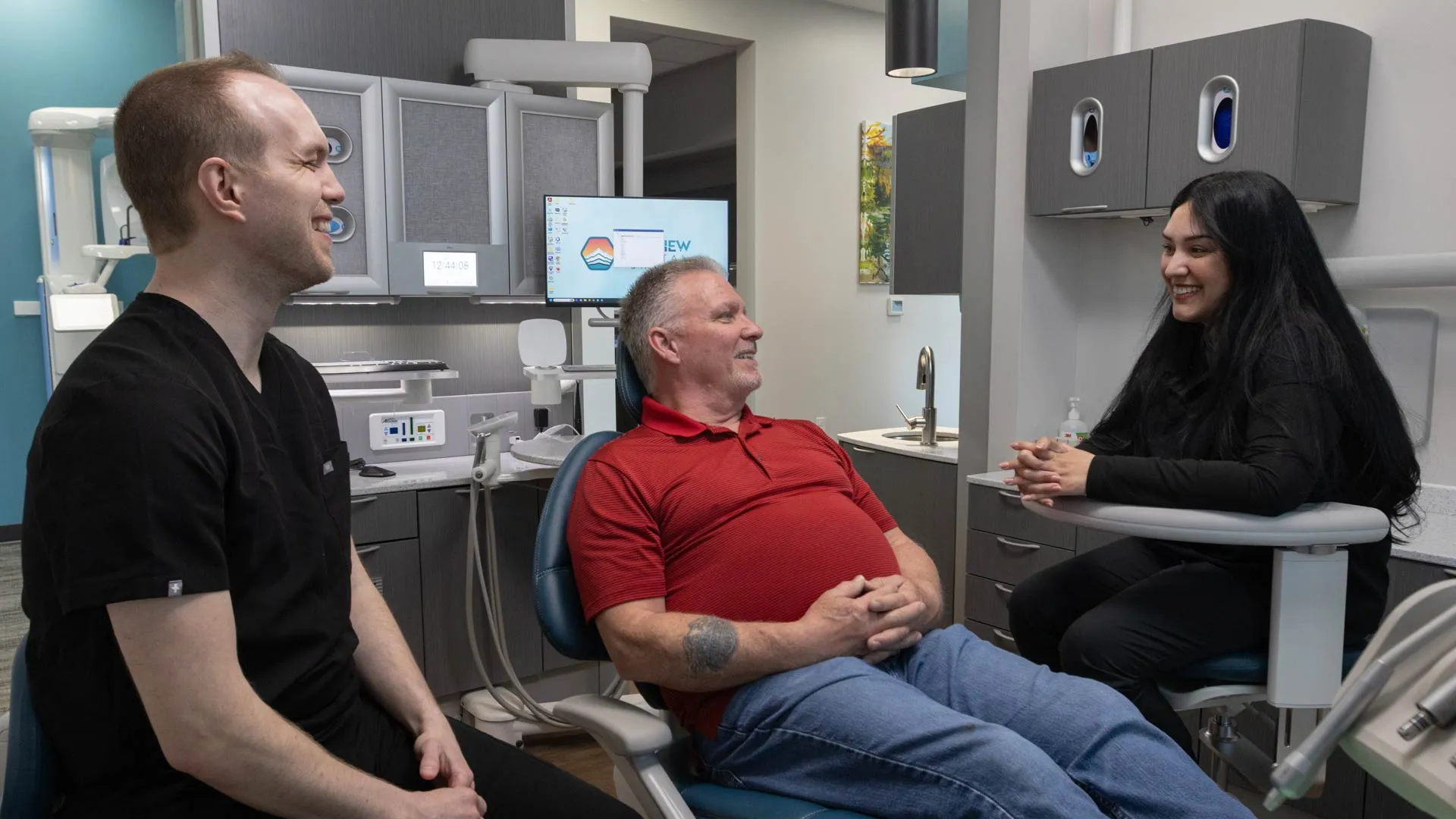 Dentist and dental assistant smiling and talking with a male patient in a modern dental office.