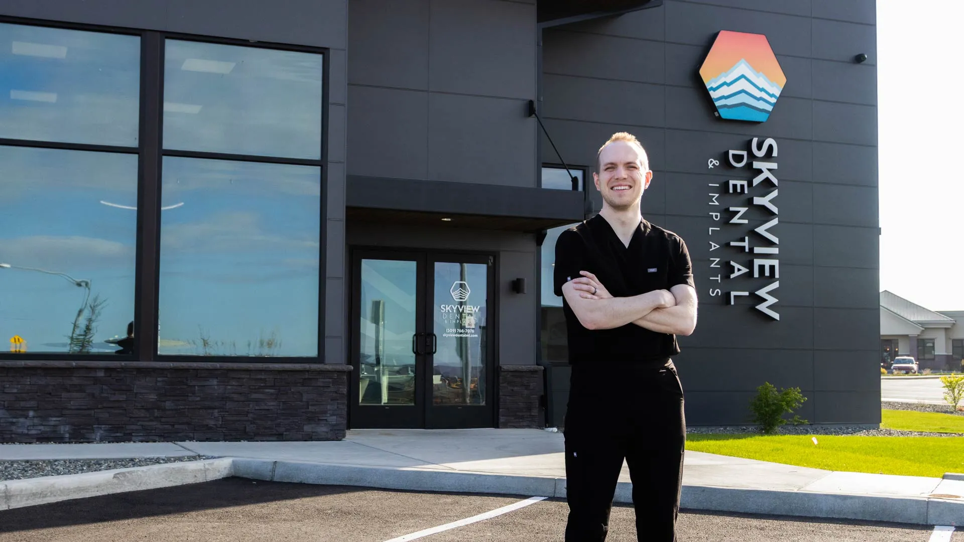 Smiling dentist in black scrubs stands outside Skyview Dental Implants clinic with arms crossed on sunny day.