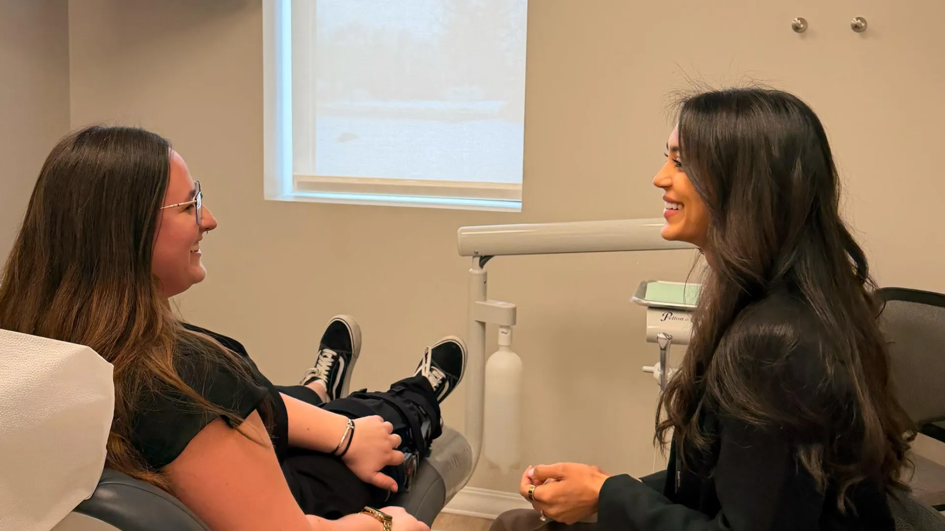 Two women smiling and talking in a modern dental office with one seated in a patient chair.