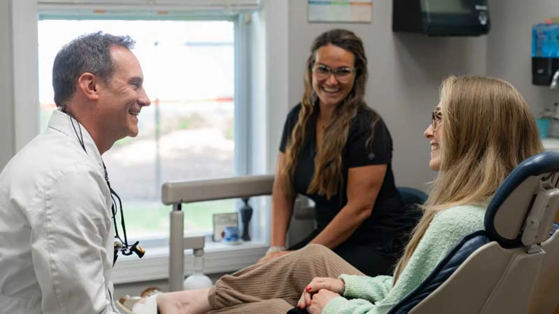 Dentist and assistant smiling and talking with a female patient seated in a dental chair by the window.