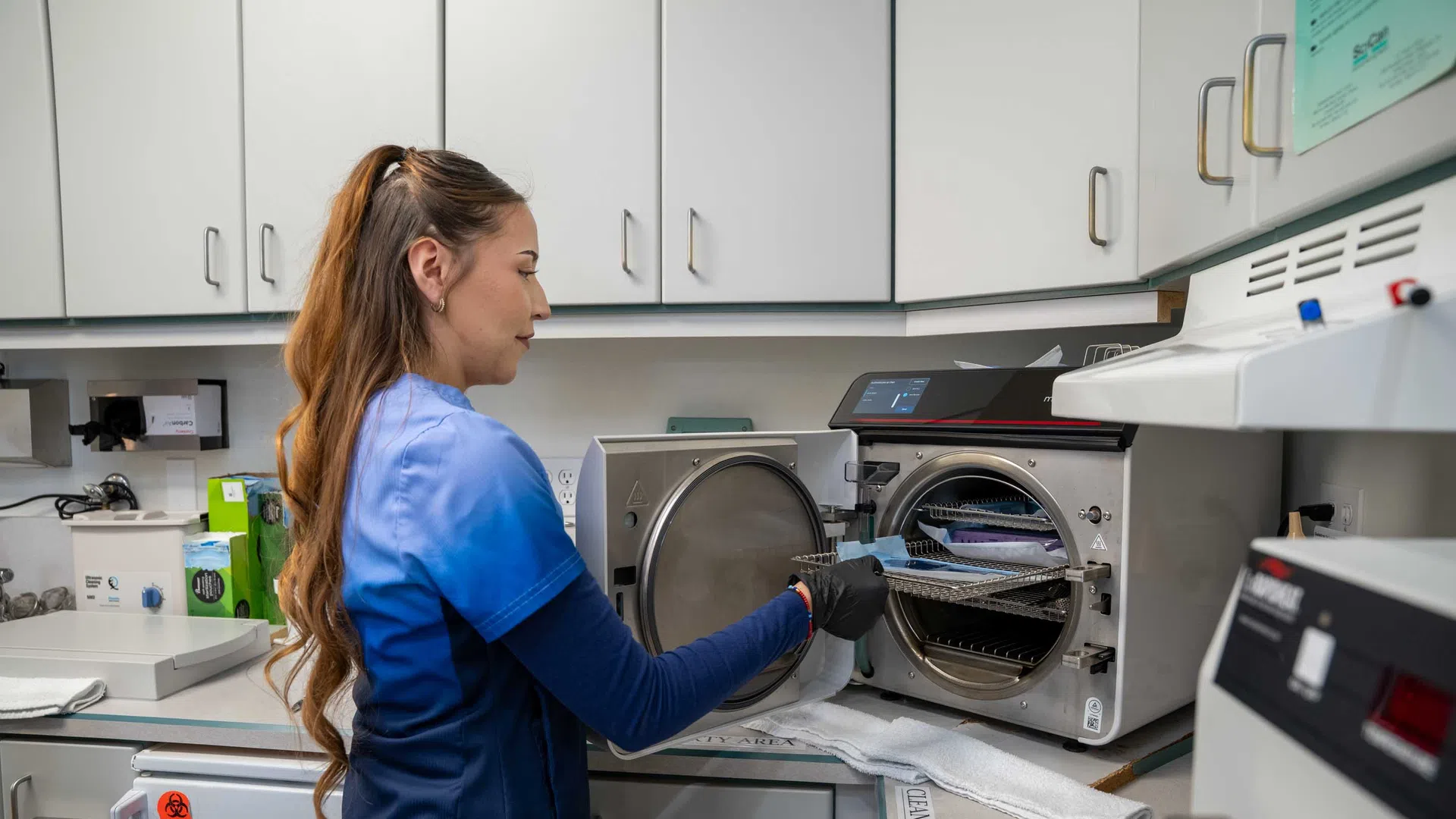 Dental professional in blue scrubs operating autoclave sterilizer in a clean, organized dental clinic workspace.