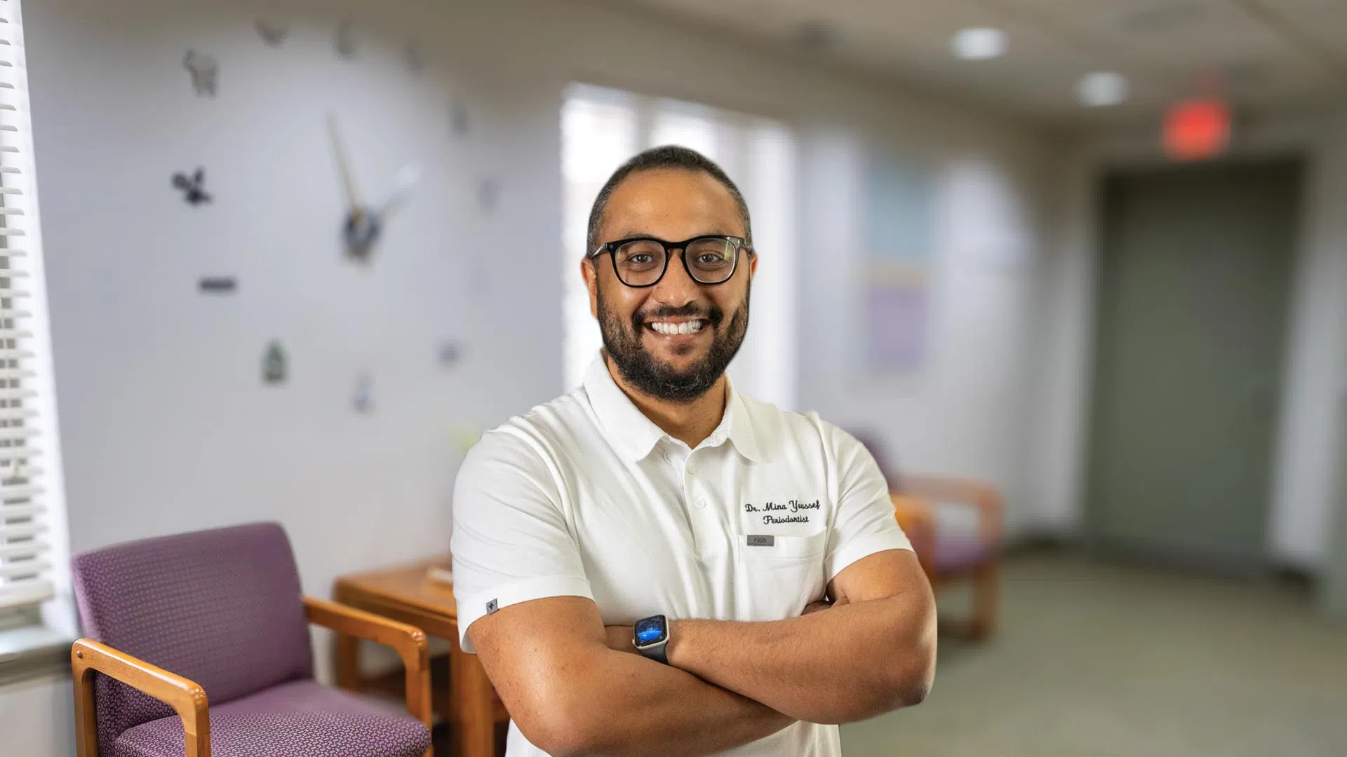 Smiling male doctor with glasses and beard stands with arms crossed in modern medical office lobby.