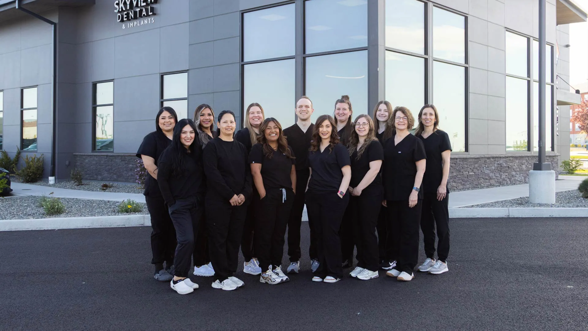 Group photo of smiling dental staff in black scrubs standing outside Skyview Dental & Implants clinic on a sunny day.