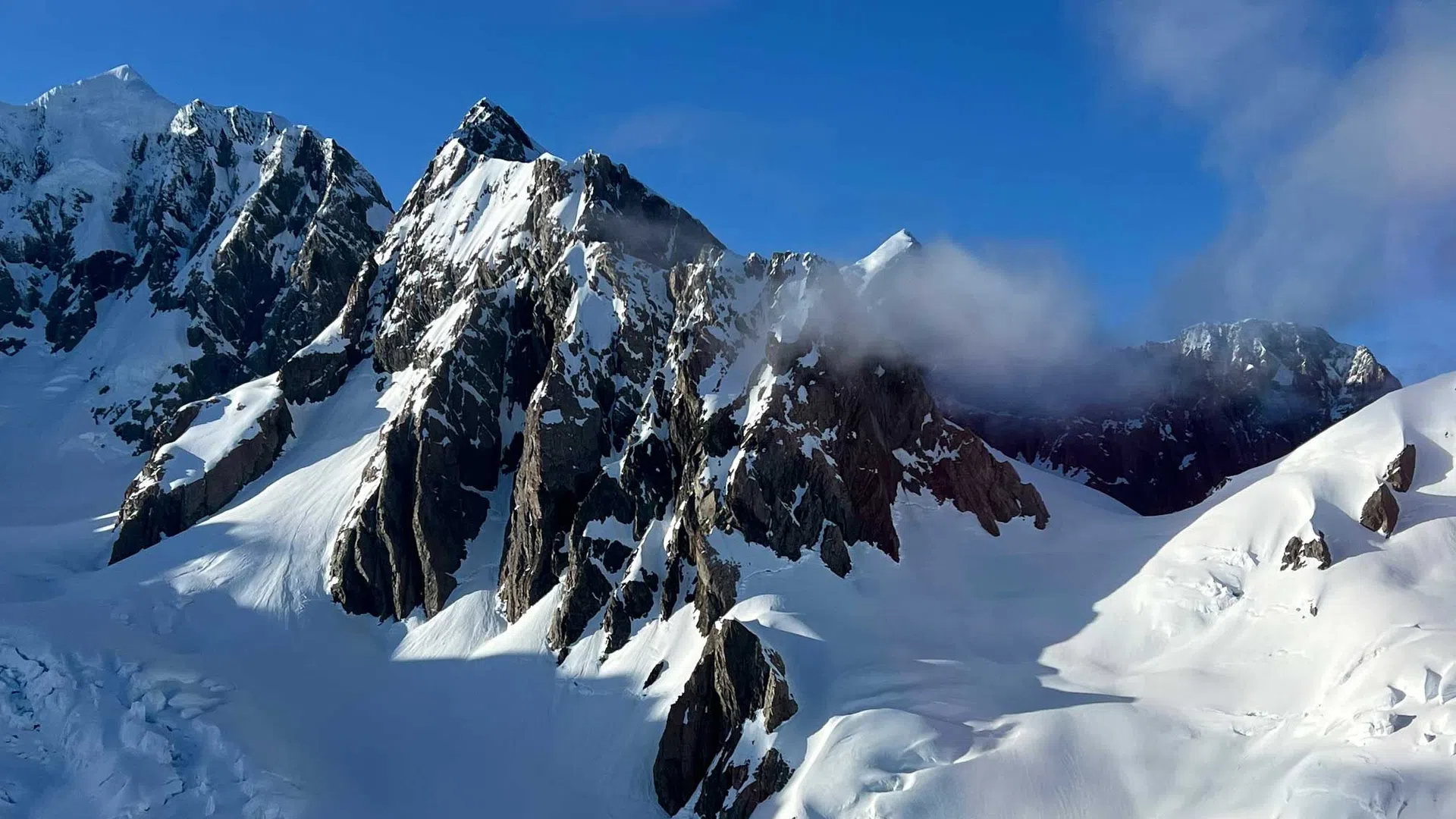 Snow-covered jagged mountain peaks under a bright blue sky with light clouds casting shadows