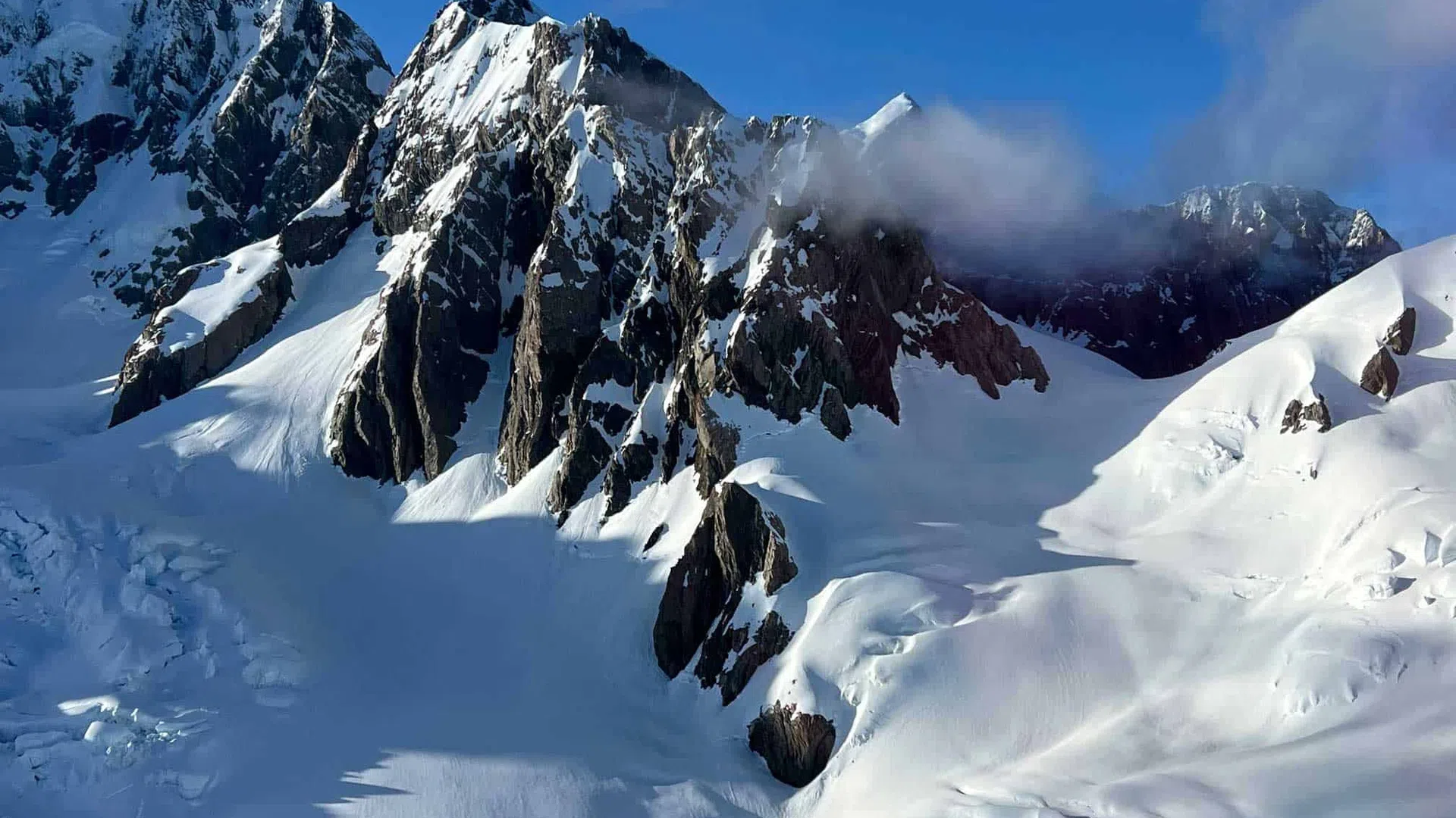 Snow-covered jagged mountain peaks under a bright blue sky with light clouds casting shadows