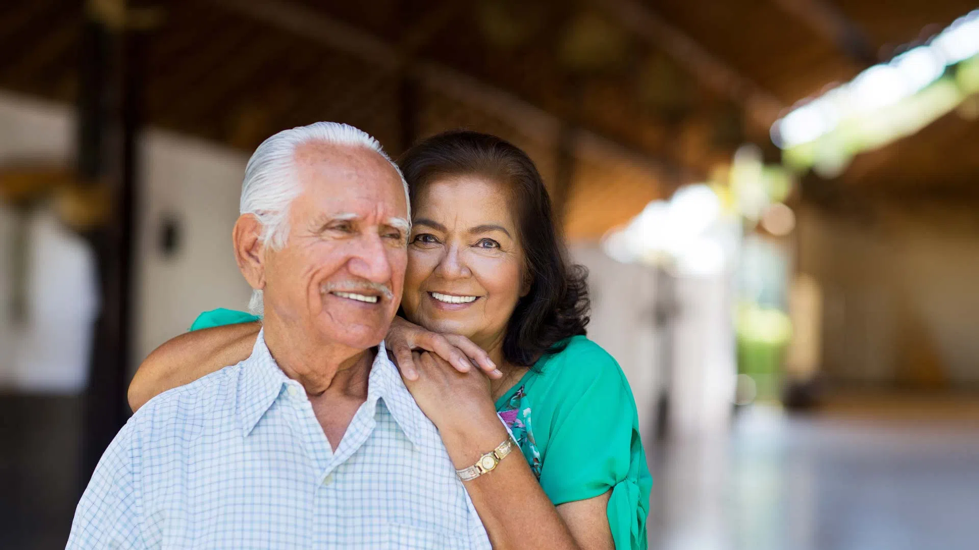 Happy elderly couple smiling and embracing in a bright, open space with wooden beams and soft lighting.