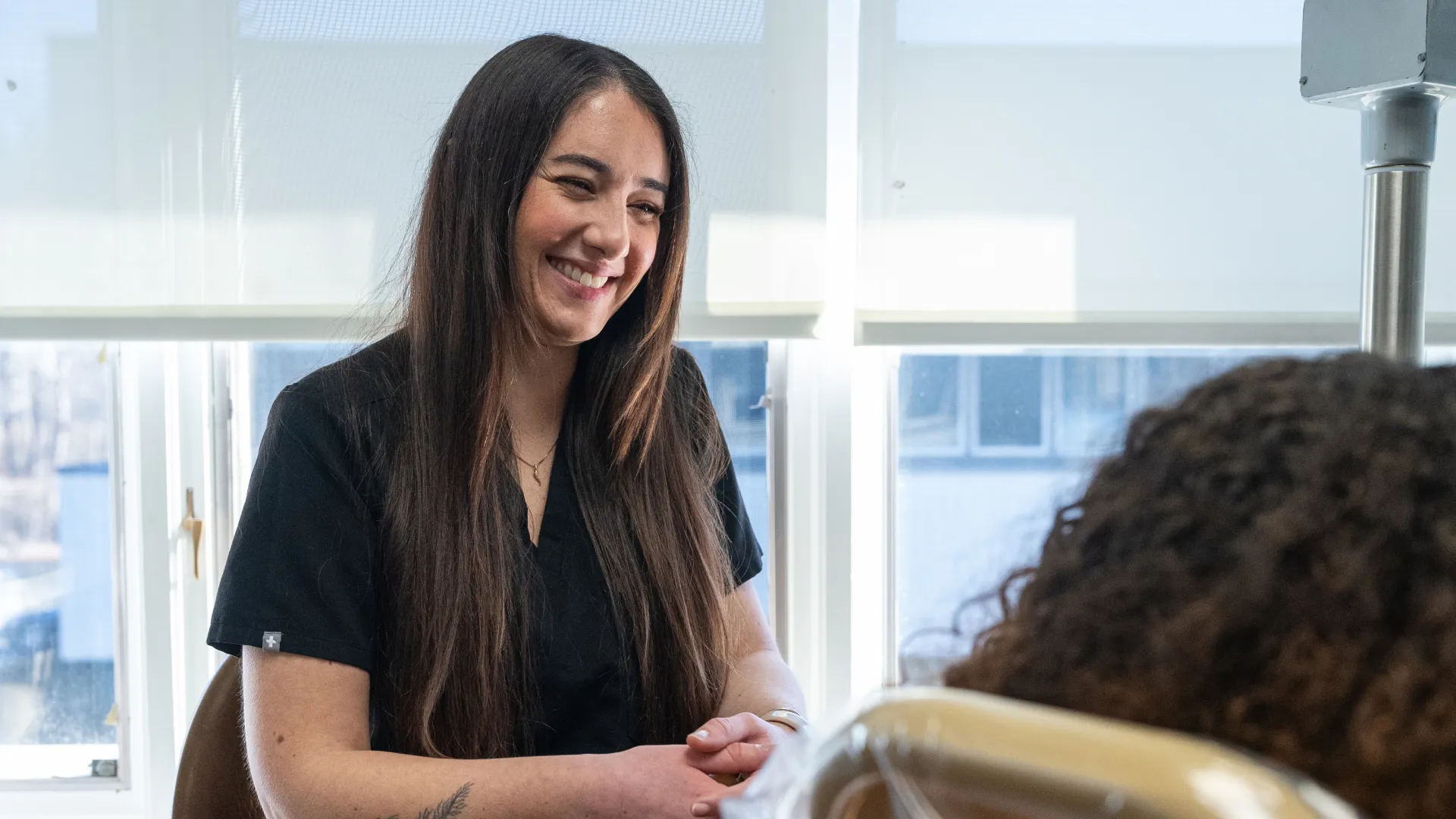 Smiling female dentist or hygienist in black scrubs sitting and talking to a patient in a dental office.
