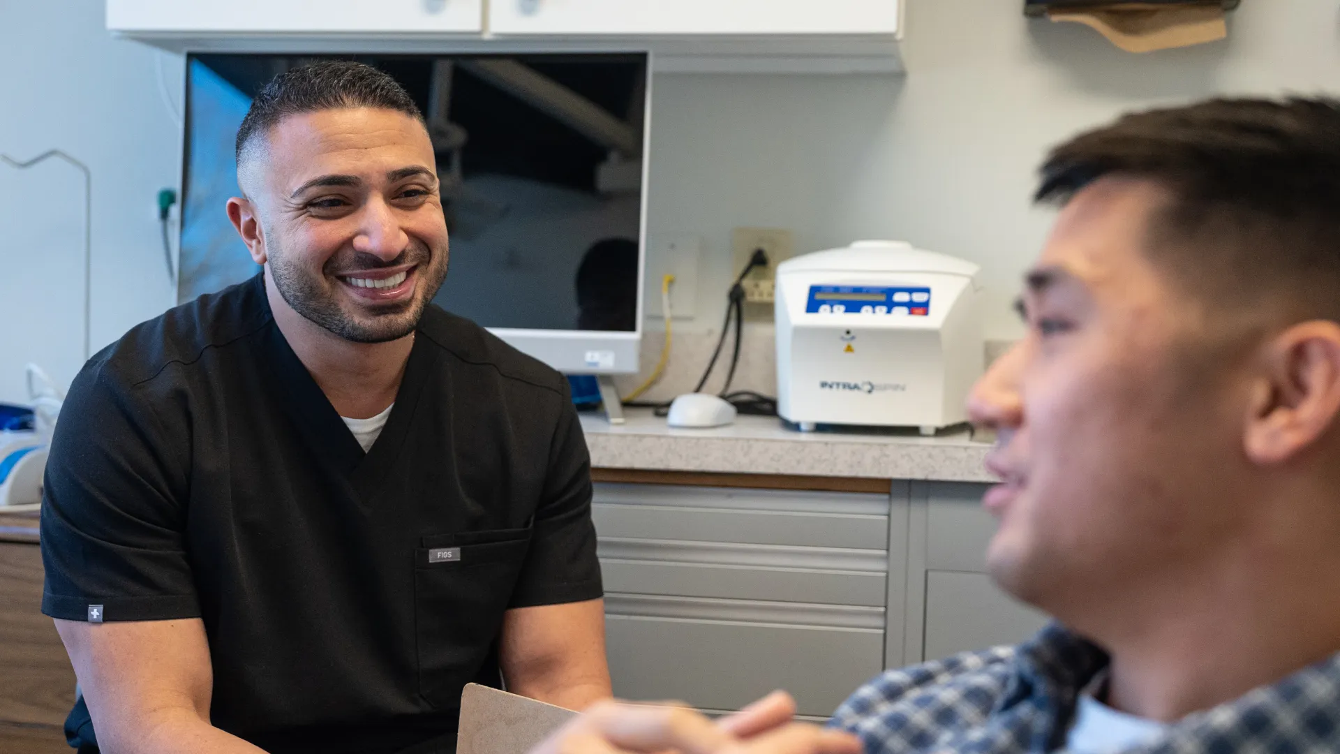 Smiling healthcare professional in black scrubs talks with patient in medical office setting.