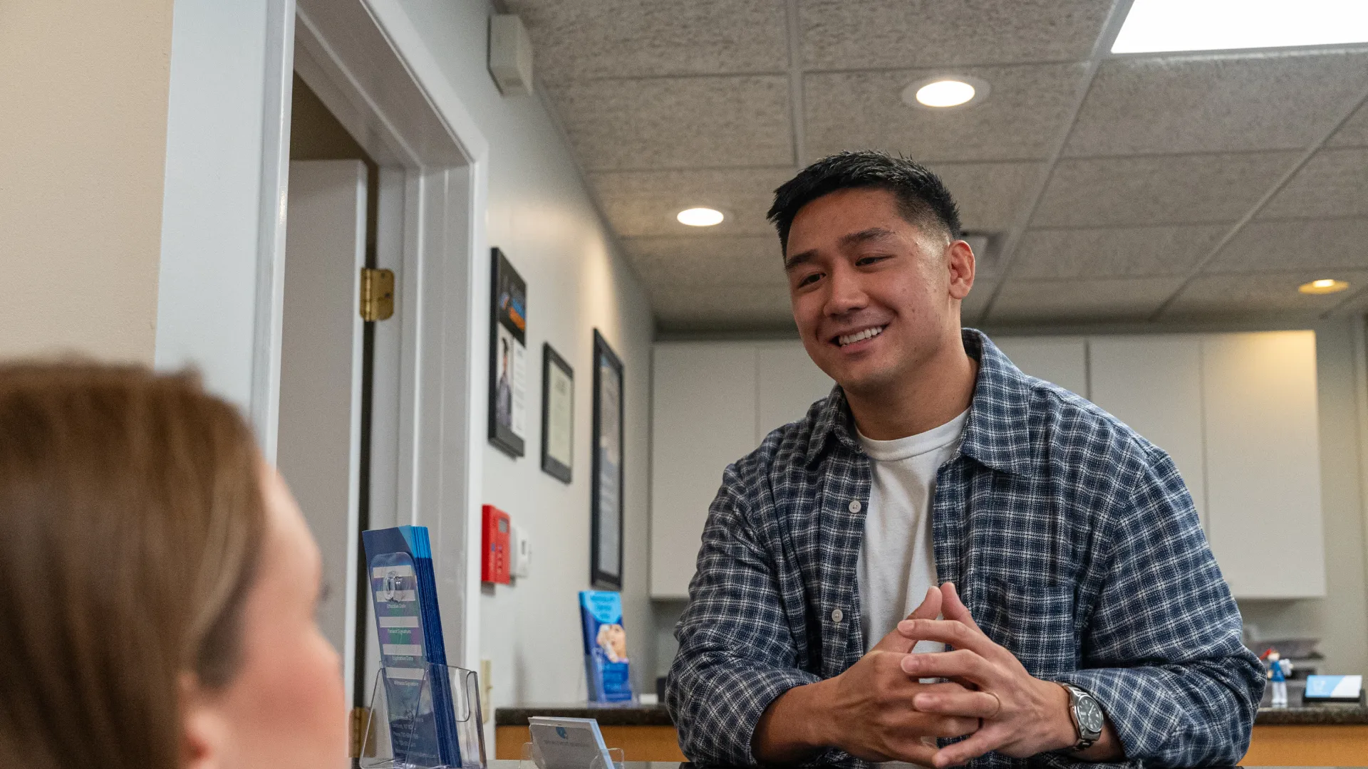 Man in plaid shirt smiles and talks with a woman at an office reception desk with informational brochures