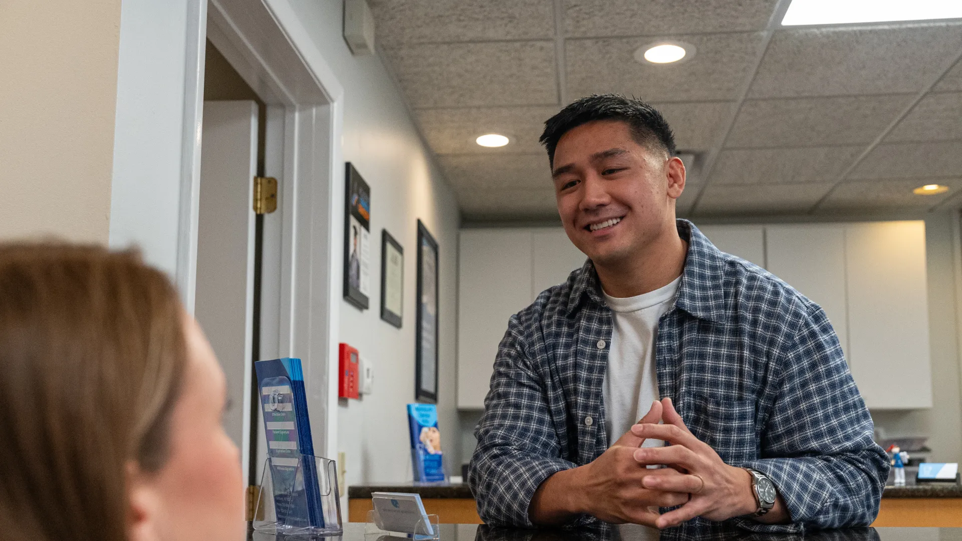 Man in plaid shirt smiles and talks with a woman at an office reception desk with informational brochures