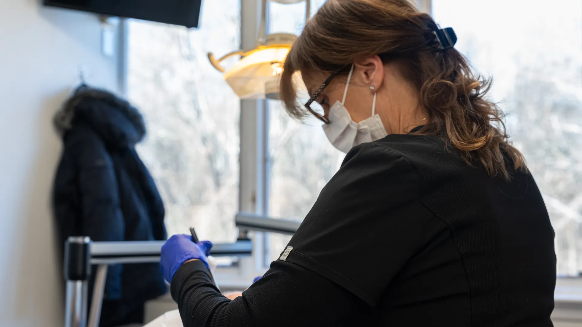 Female dentist wearing mask and gloves working on a patient in a bright dental clinic with large windows.