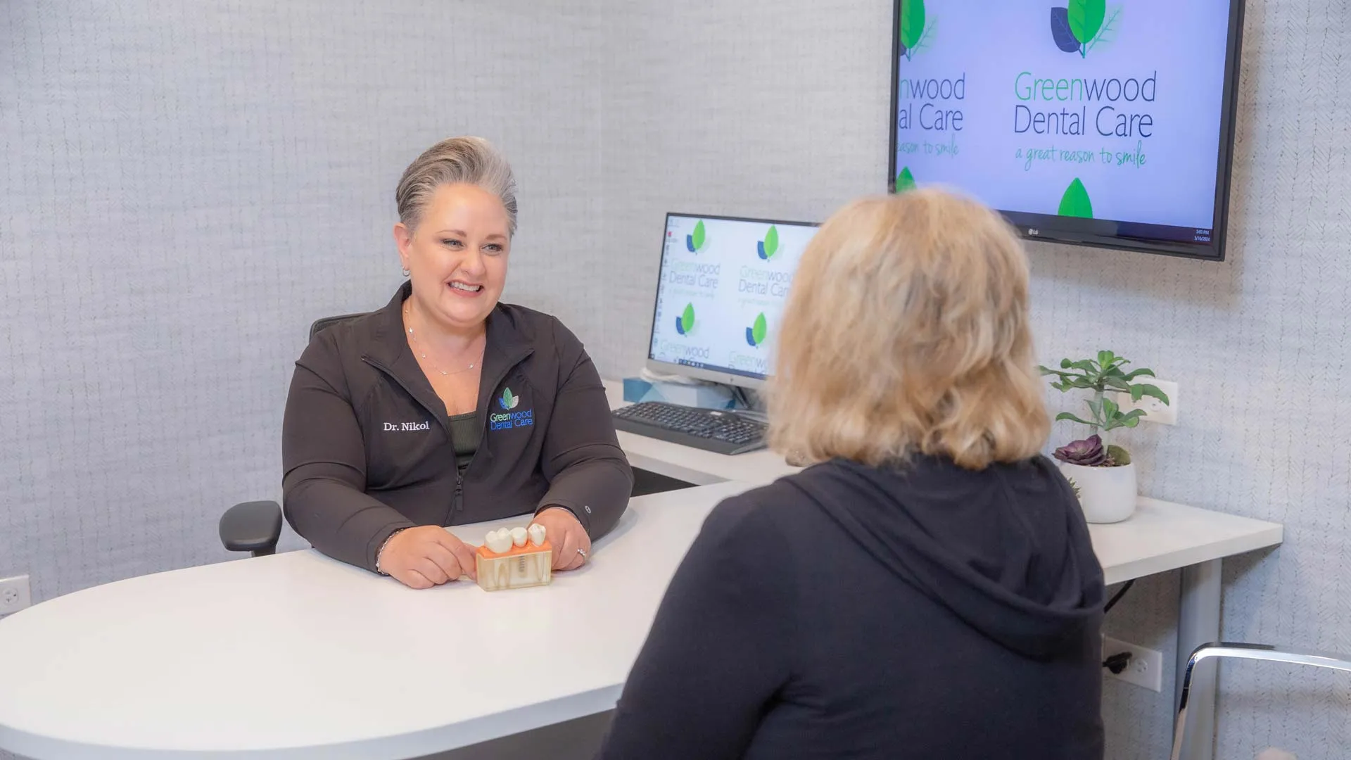 Dentist in uniform explaining dental care using tooth model to a patient in modern consultation room.