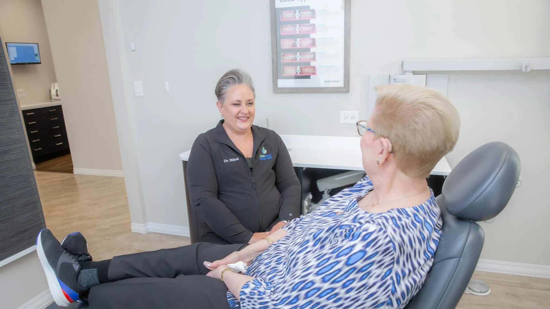 Female patient sitting in dental chair talking with female dentist in modern clinic setting.