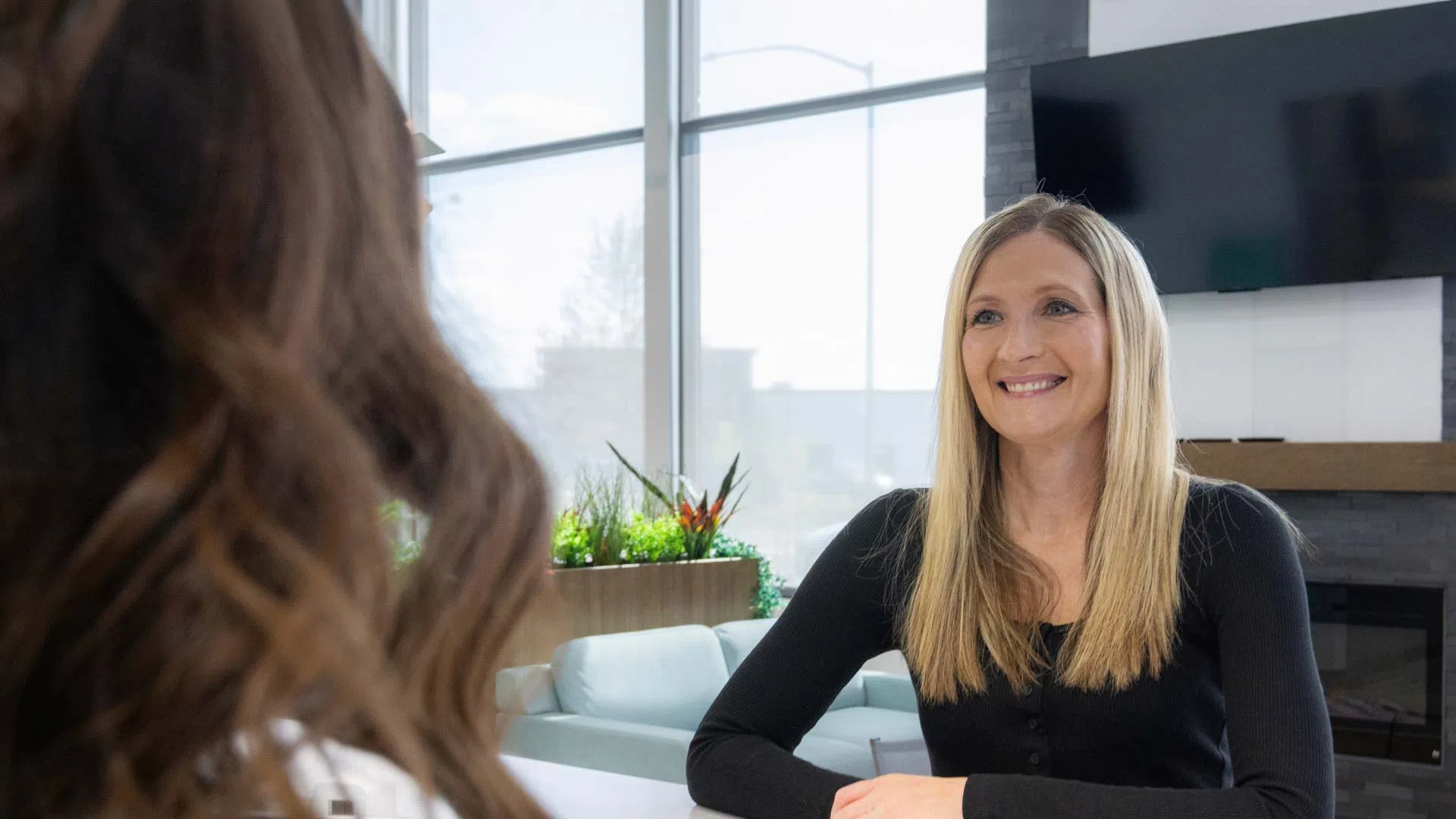 Two women engaged in a friendly conversation in a modern office with large windows and natural light.