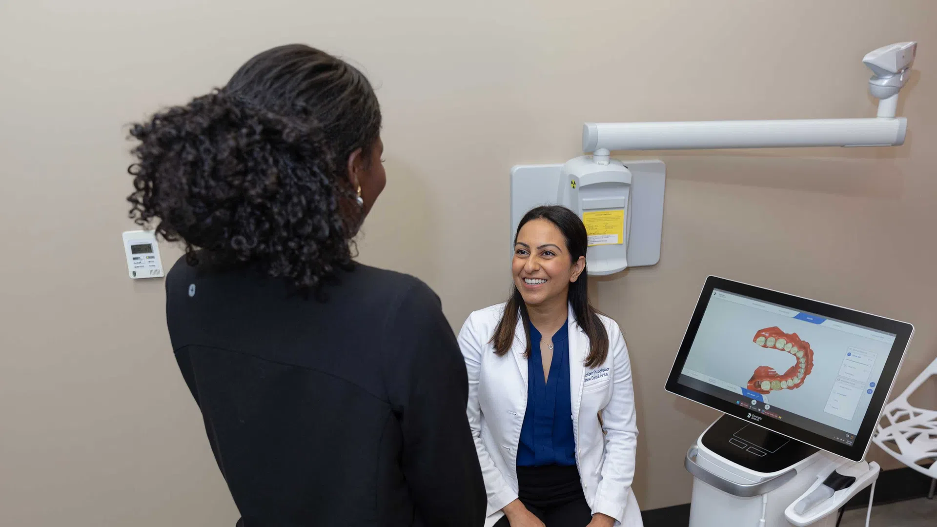 Dentist consulting with patient next to digital dental imaging equipment displaying teeth scan.
