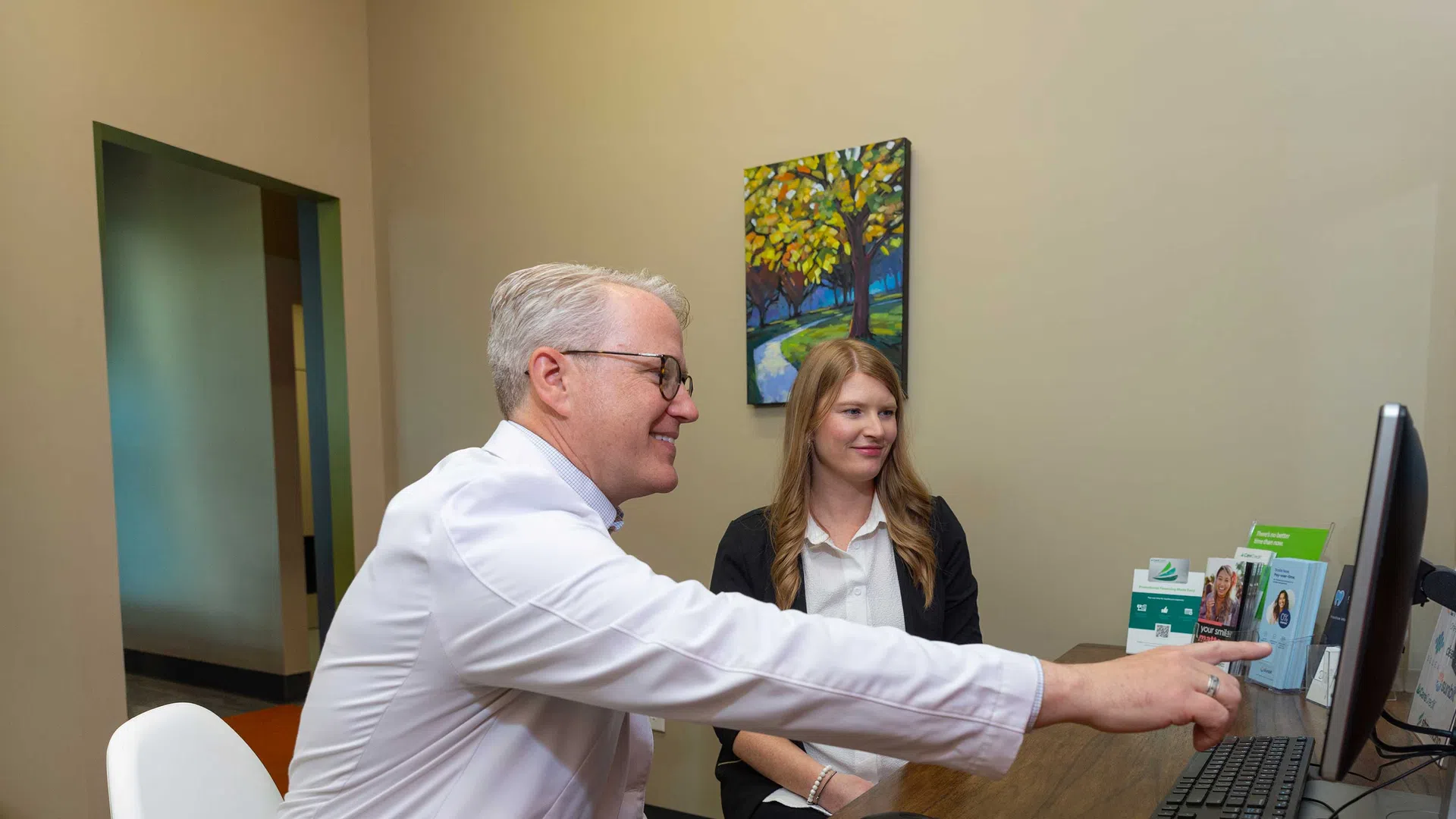 Doctor explains health information on computer screen to smiling female patient in consultation room.