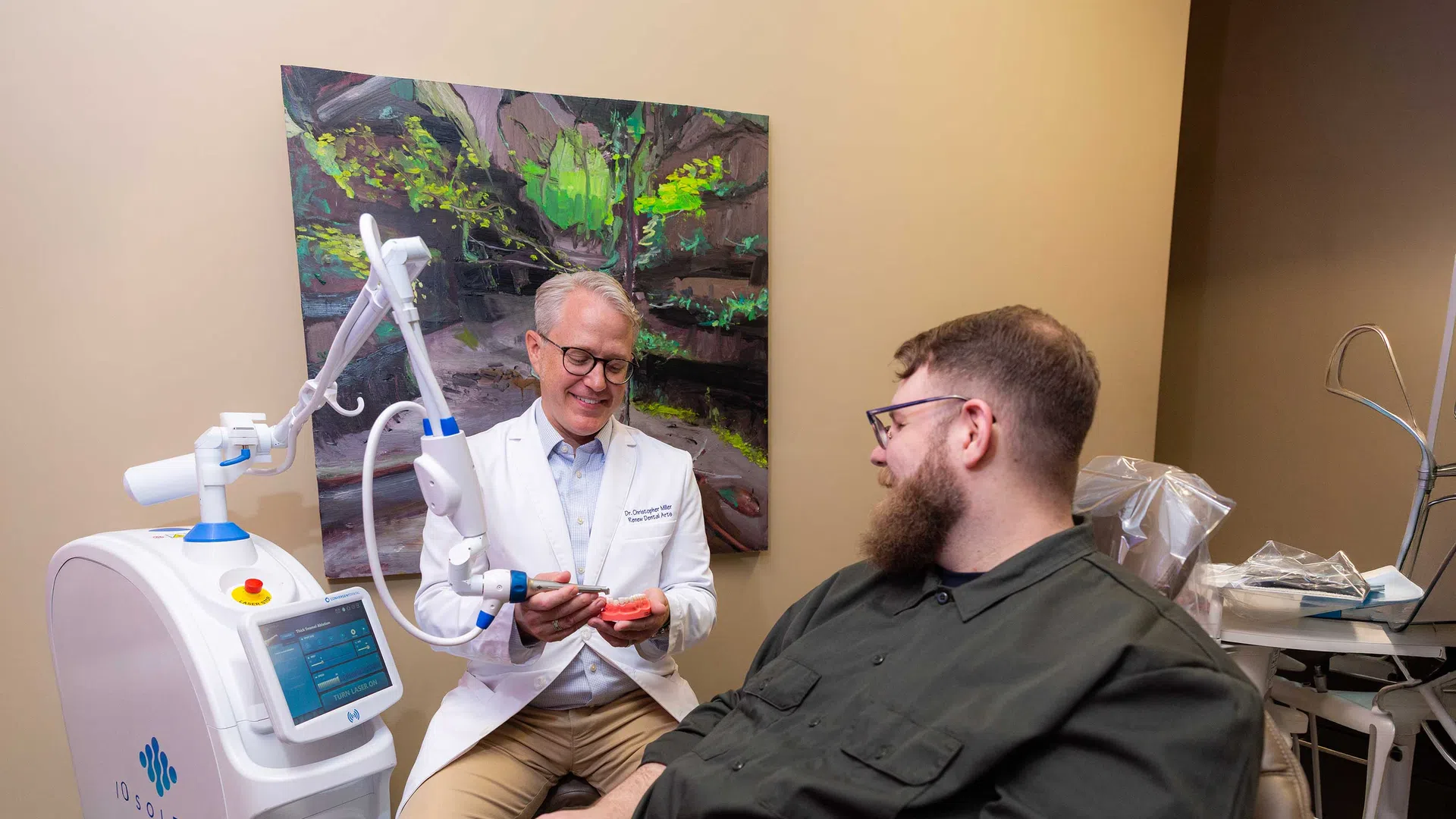 Dentist demonstrating dental laser treatment on a model while patient watches in a dental office.