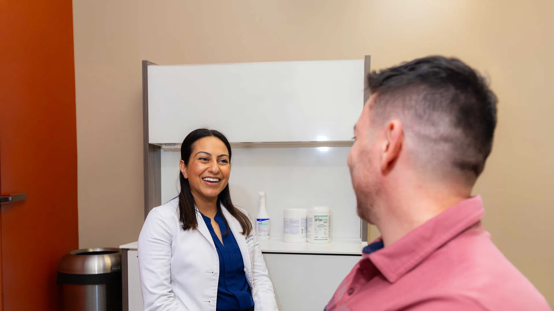 Female doctor in white coat smiling and talking to male patient in clinical office setting.