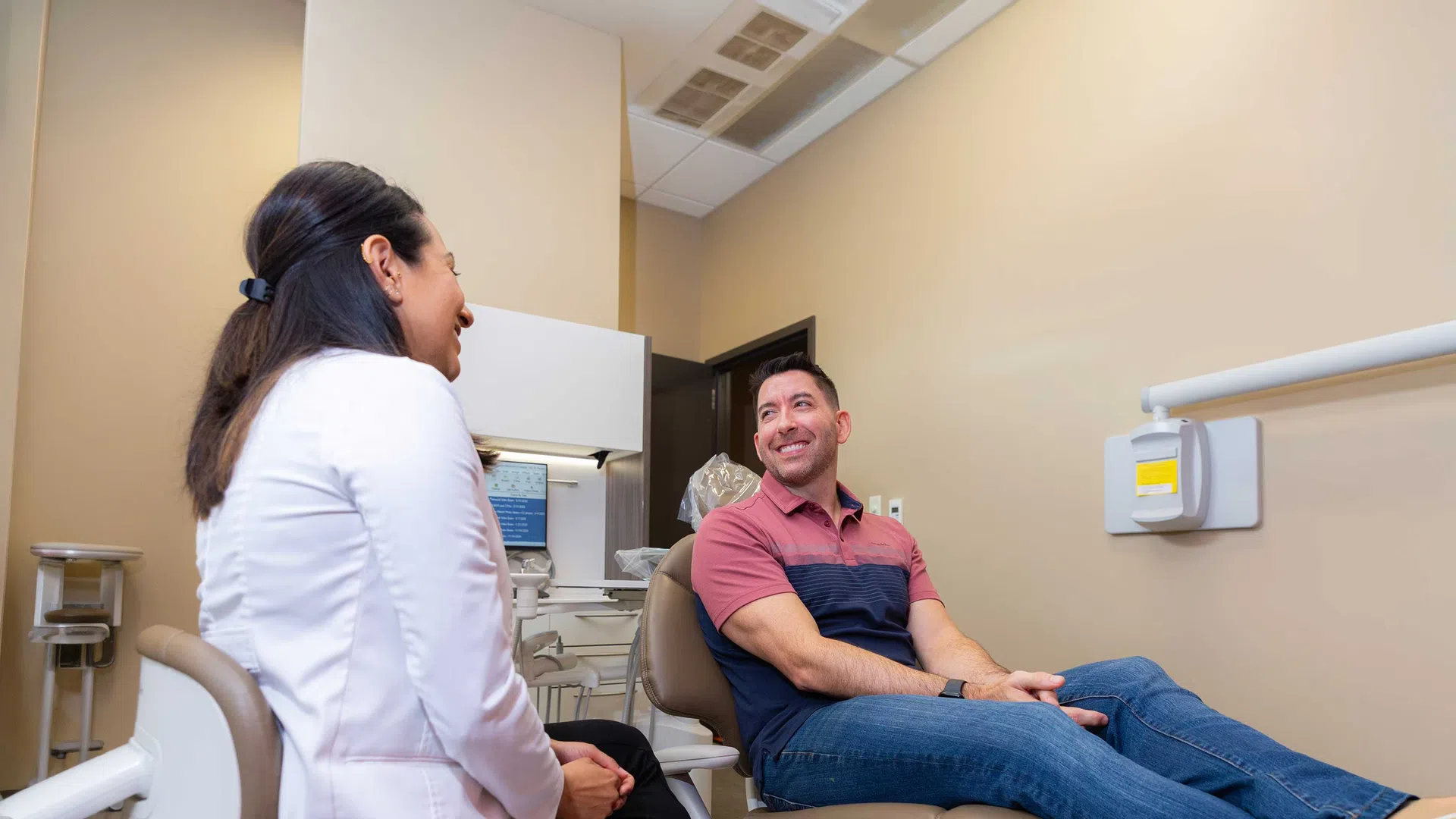 Dentist and male patient smiling and talking in a modern dental exam room.