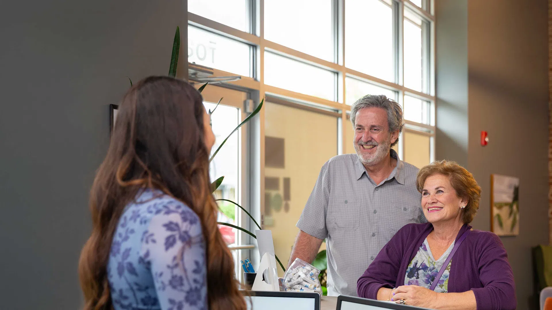 Dental clinic receptionist assisting elderly couple at front desk with computer screens displaying Renew Dentistry logo.