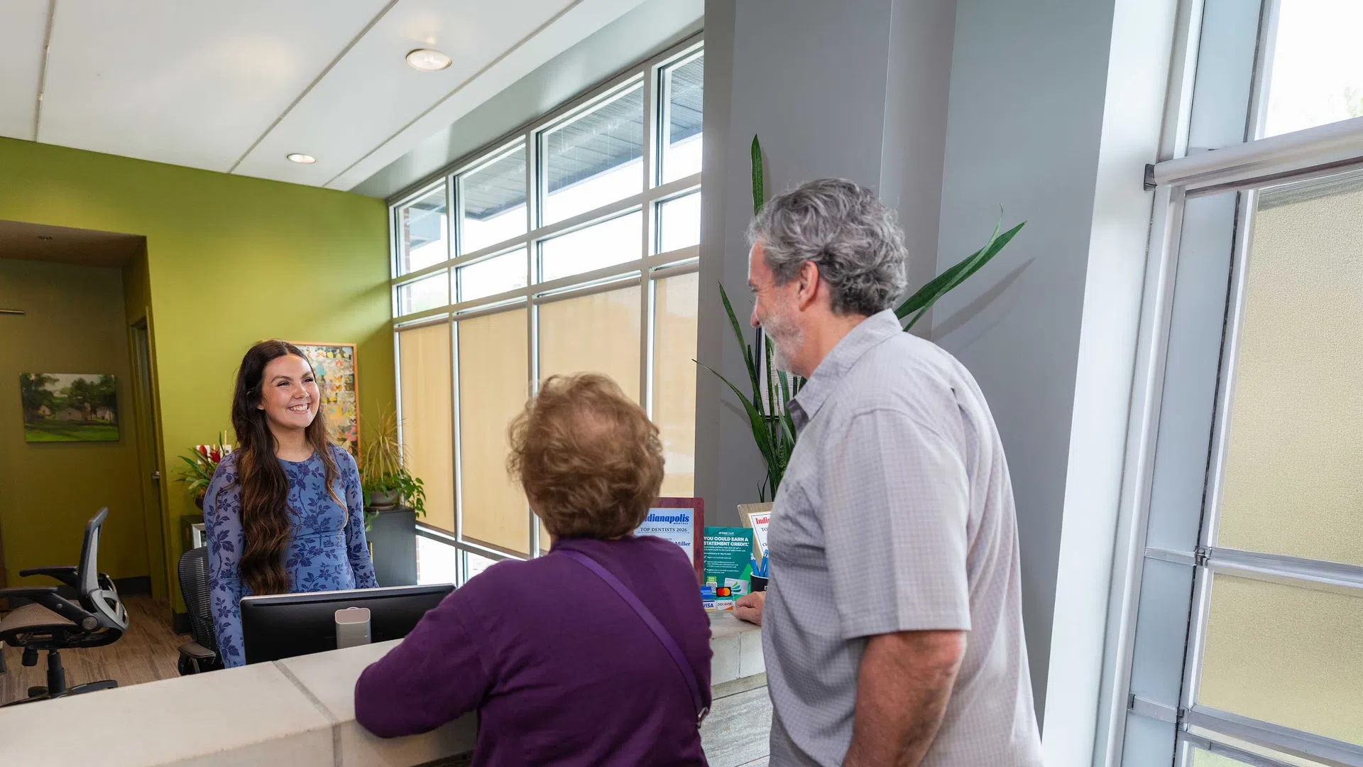 Smiling receptionist assisting elderly couple at modern clinic reception desk with bright windows and plant