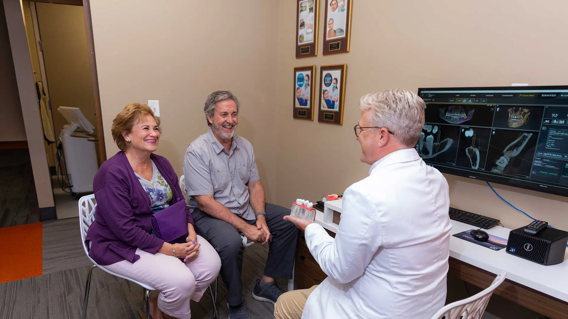 Dentist explaining dental model to smiling senior couple in consultation room with 3D dental images on screen
