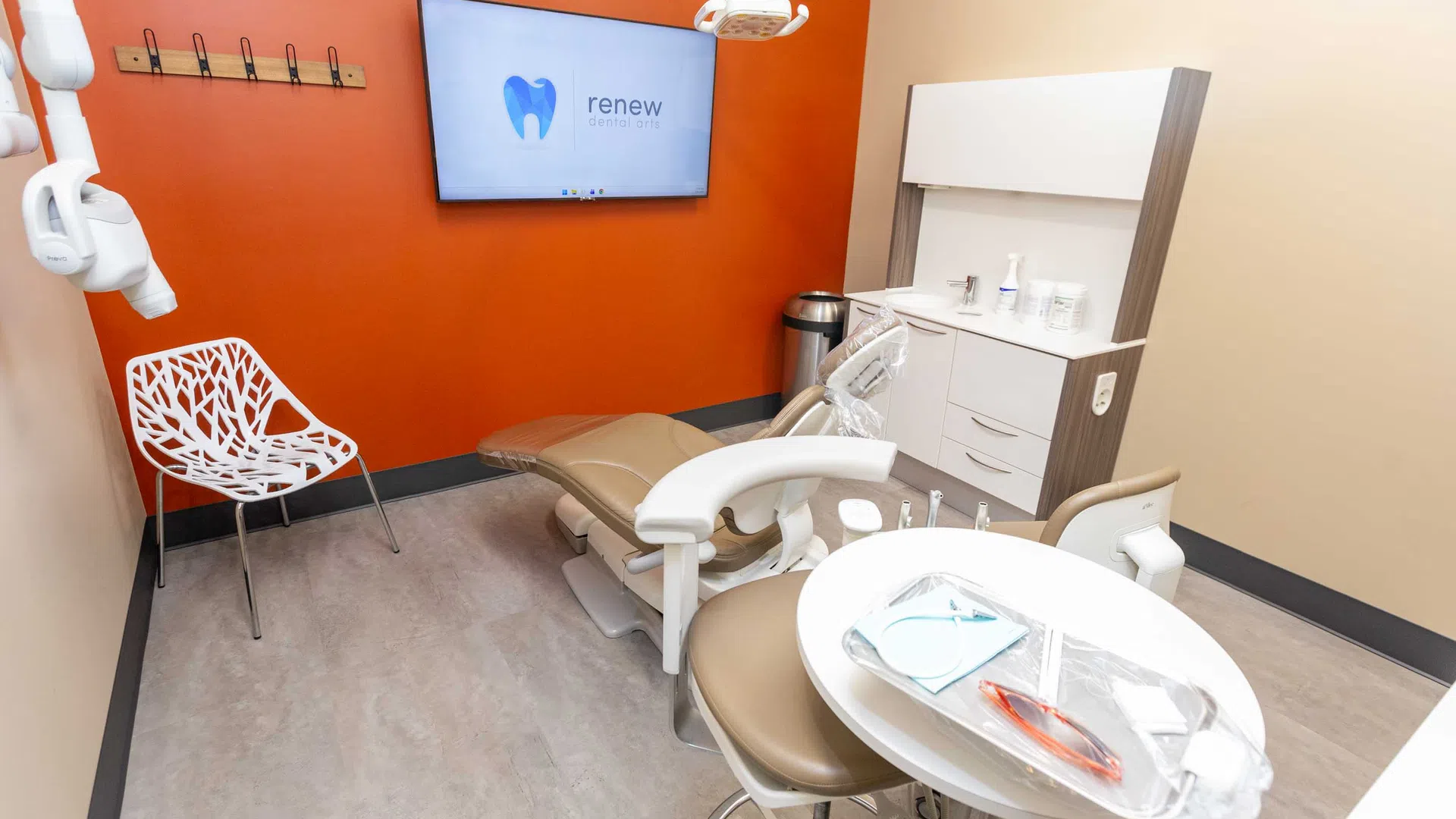 Modern dental office with beige chair, orange accent wall, white shelving, and screen displaying Renew Dental Arts logo.