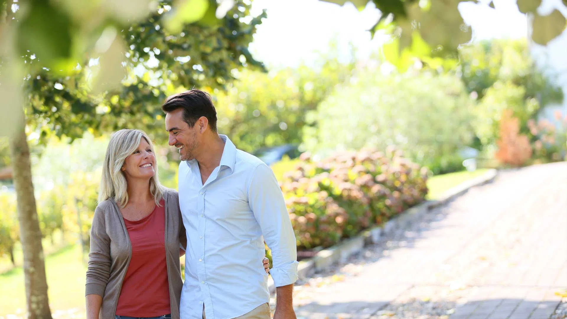 Smiling couple walking arm in arm on a sunlit garden pathway surrounded by green trees and flowering bushes