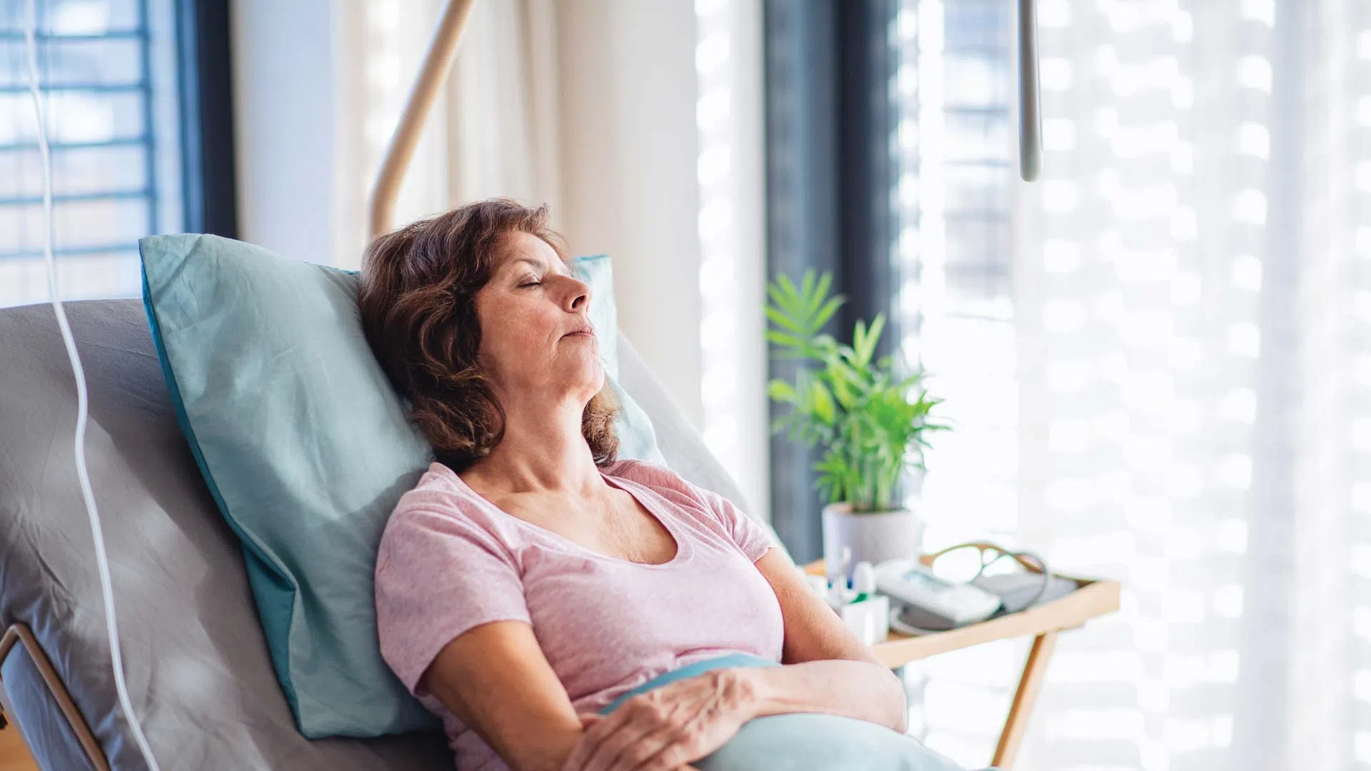 Middle-aged woman resting in hospital bed with eyes closed in a bright patient room with medical equipment.