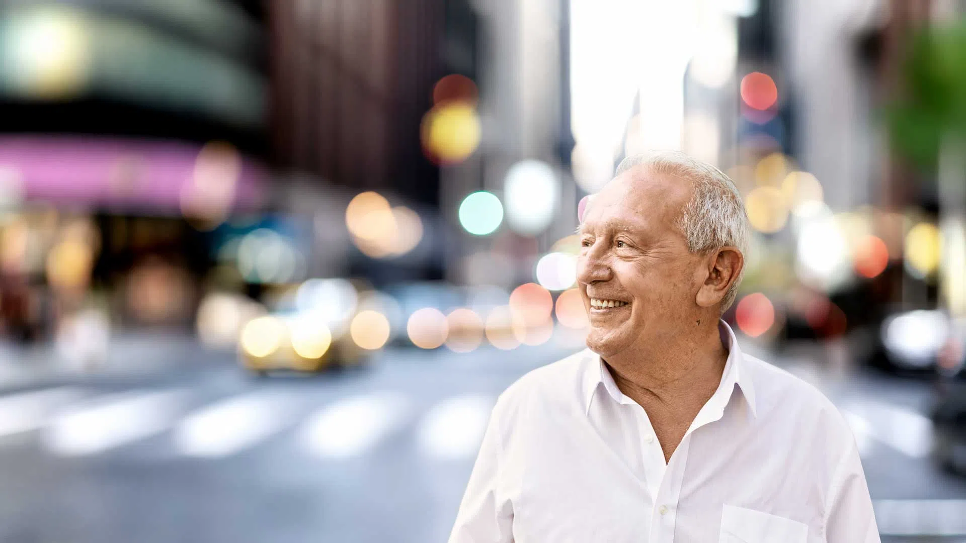 Smiling elderly man in white shirt standing on a city street with blurred urban lights in background