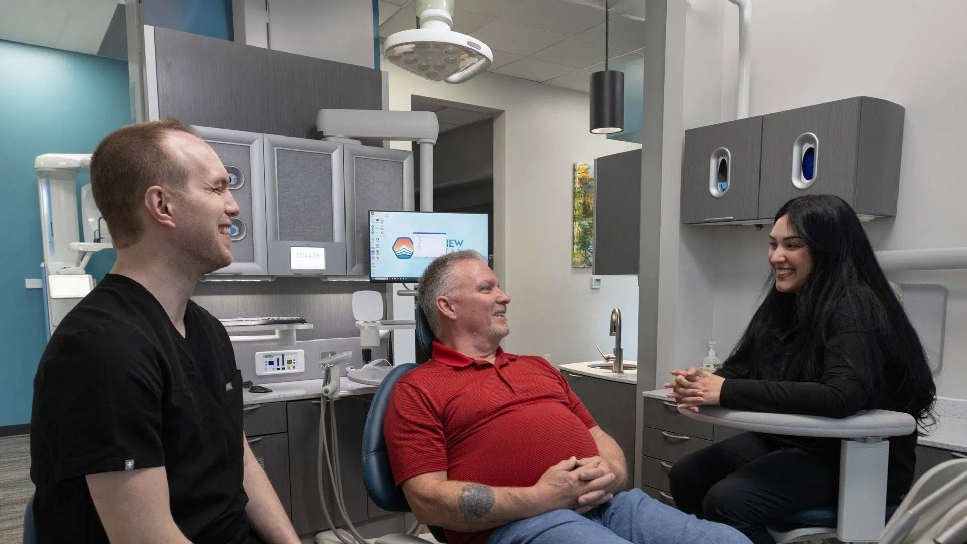 Dentist and two adults smiling and talking in a dental office with modern equipment and cabinetry.