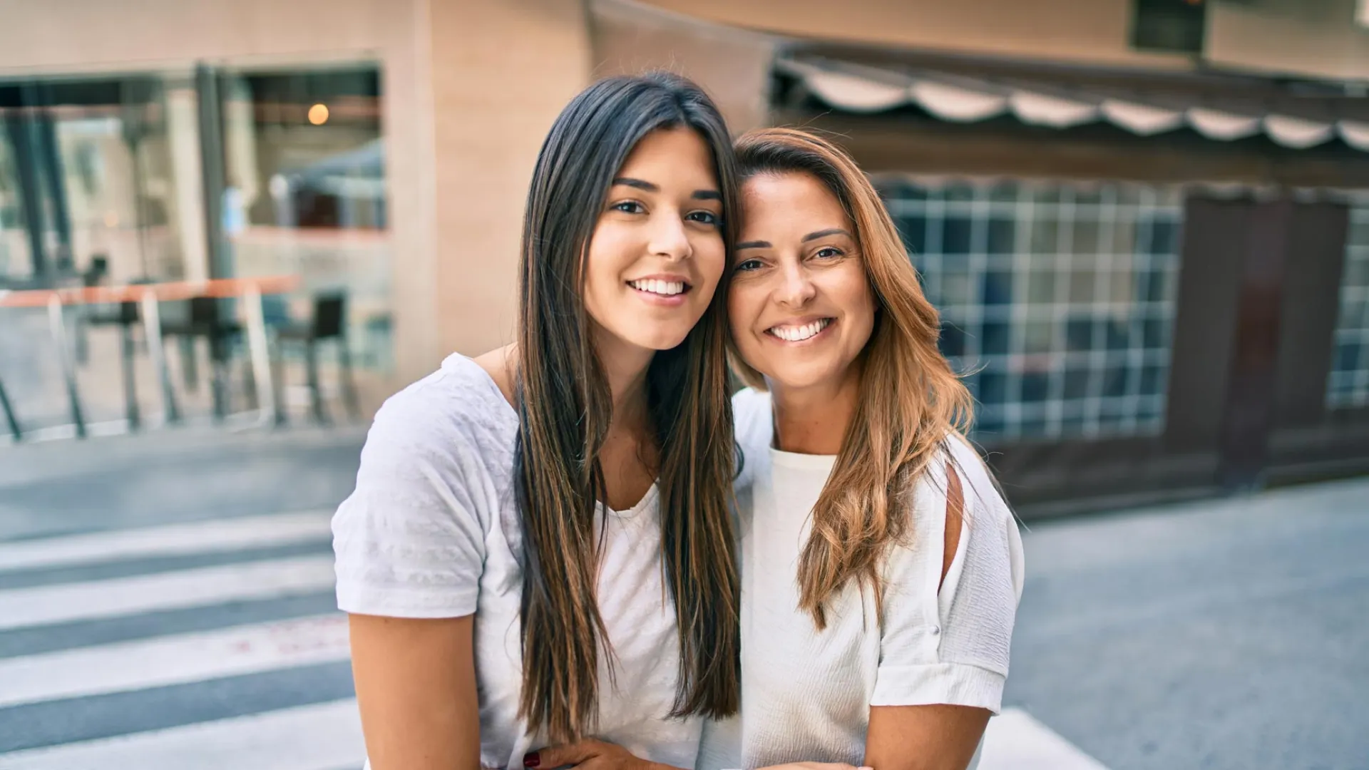 Two smiling women standing close together outdoors, posing for a happy portrait on a city street.