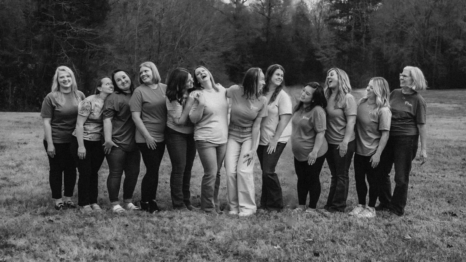 Black and white photo of eleven women standing in a field, smiling and interacting happily in casual attire.